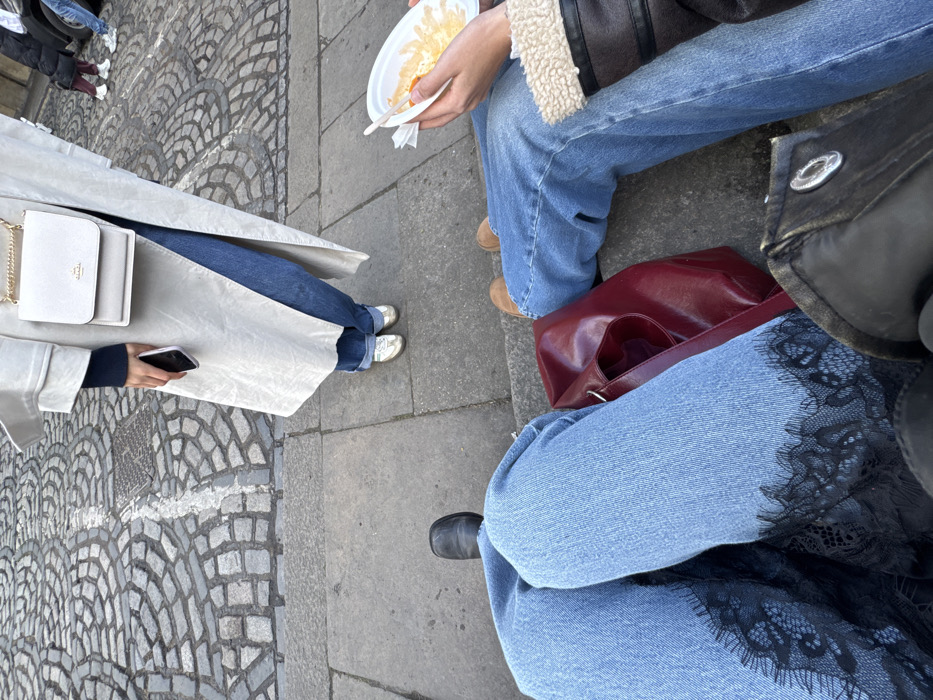 Three people are seated outdoors on a concrete surface. One person wears a light-colored trench coat, jeans, and metallic sneakers, holding a phone. Another person wears a shearling-lined coat, jeans, and tan shoes, holding a plate with food. The third person wears jeans with black lace trim and black shoes, with a red purse and a black bag nearby. The surface under them is a mix of concrete and cobblestones.