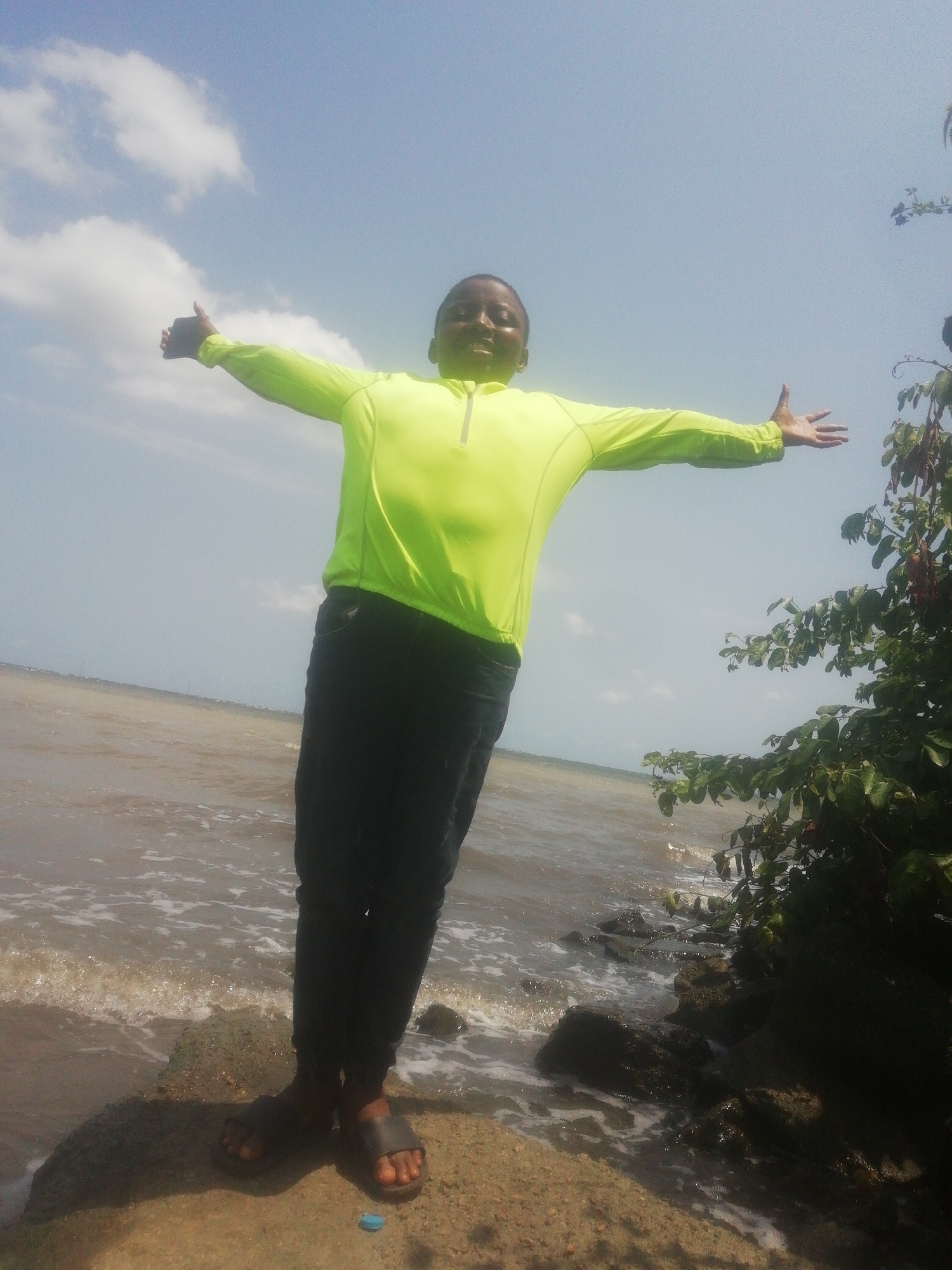 A person stands on a rock at the water's edge with arms outstretched. The person wears a bright green long-sleeved shirt, dark pants, and sandals. The water appears murky and choppy. In the background, the sky is partly cloudy. Some greenery is visible to the right of the person. The location is Unknown, Unknown.