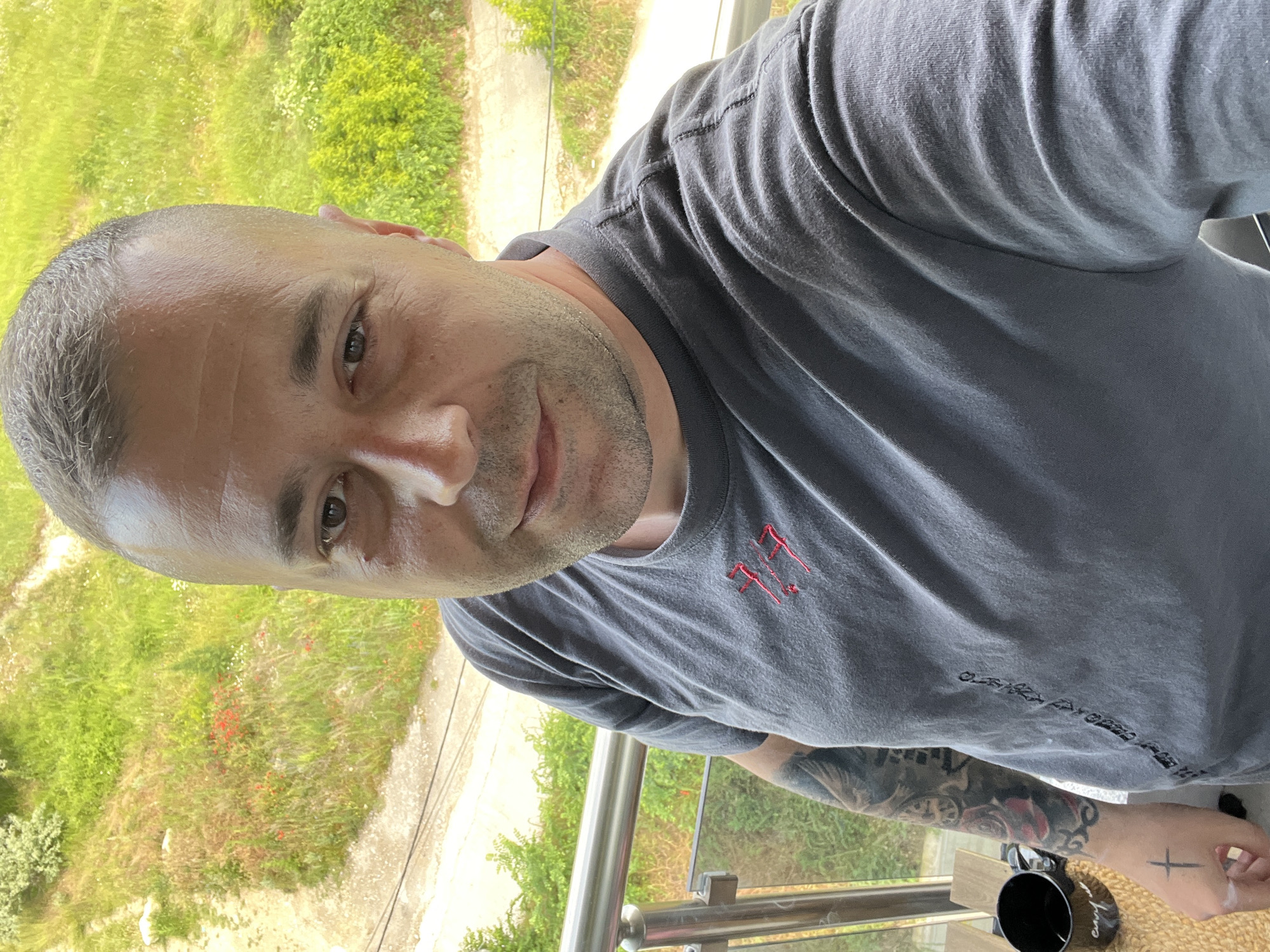 A man with short, buzzed, graying hair and stubble on his face is taking a selfie on a balcony. He is wearing a dark gray t-shirt with red embroidery on the left side of the chest and a tattoo on his left arm. A black mug is visible on the right side of the frame, with a hand reaching towards it. Behind him, a railing and glass partition are visible, suggesting he is on an upper floor. Beyond the balcony, lush green foliage and a dirt path are seen, indicating an outdoor, possibly semi-rural, setting. The lighting suggests it is daytime. The image is cropped at the top, cutting off the top of the man's head and the background. The image was taken on a sunny day in Varna, Bulgaria.