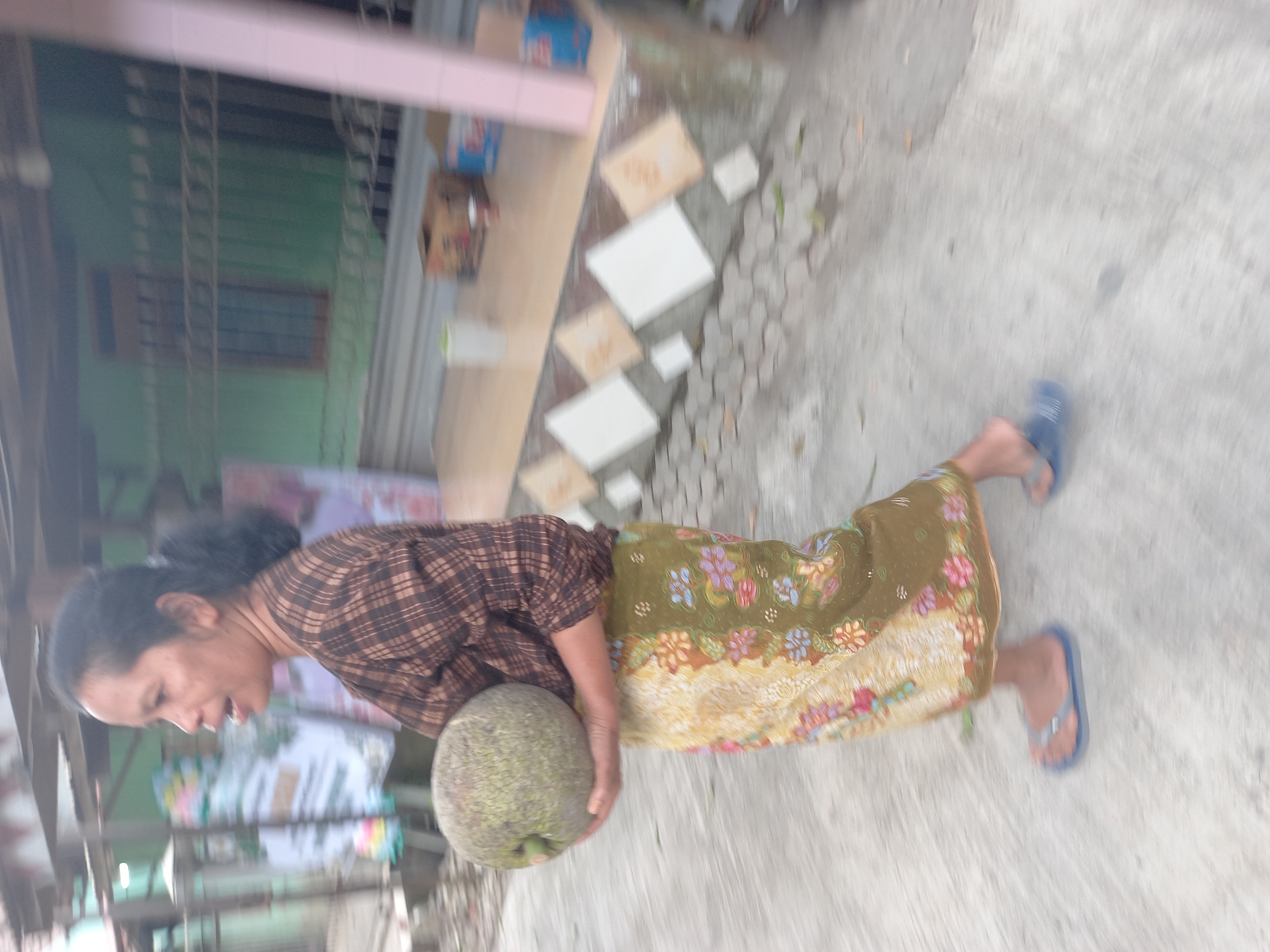 A woman walks outdoors in Banyumulek, Indonesia, carrying a large, green, spiky fruit. She is wearing a brown plaid long-sleeved shirt and a long, patterned skirt in shades of green, yellow, and red. She has dark hair pulled back and is wearing blue flip-flops. The ground appears to be concrete. In the background, there is a building with green walls and a fence, and a light pink pole is visible on the right side of the frame. The scene suggests a residential or commercial area.