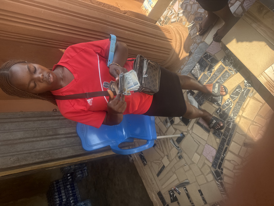 A female individual stands outdoors in Aguata, Nigeria, focusing on handling currency. She is seen from the chest up, with her head slightly bowed, concentrating on a stack of Nigerian Naira banknotes in her hands, including a visible 1000 Naira note. She wears a red Adidas short-sleeved shirt, a black skirt, and black "CELINE" branded slide sandals. A dark brown shoulder bag with a repeating pattern is slung across her body. A light blue mobile phone is held or tucked under her right arm. The ground beneath her is a mosaic-style tiled surface composed of irregular beige, black, and white pieces. To her left, a blue plastic chair sits near an open dark brown doorway, beyond which numerous stacks of bottled water are visible, suggesting a retail or storage space. To her right, a light brown fluted architectural pillar is present. The lower legs and feet of a second individual, wearing dark flip-flops, are partially visible in the far right foreground. The scene is illuminated by bright natural light, casting distinct shadows.