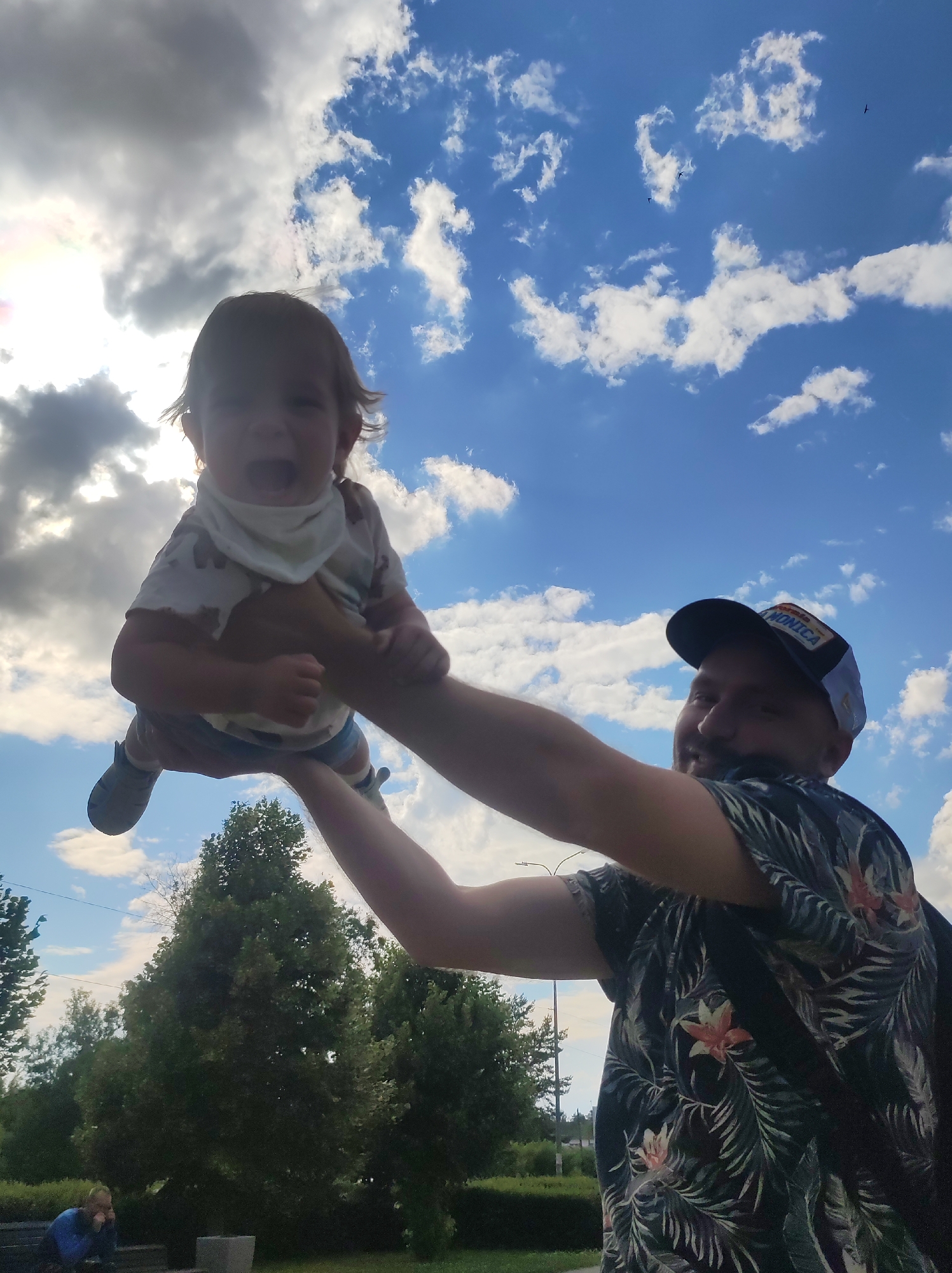 A man in a dark, tropical-print t-shirt and a baseball cap is holding a young child up in the air against a bright blue sky with scattered white clouds. The sun is shining, creating a lens flare effect. The child is wearing a light-colored t-shirt with a pattern and a bib or bandana. The child's mouth is open, possibly in a cry or a laugh. The man is smiling, looking at the child. In the background, there are trees and what appears to be a park or outdoor area. The man is wearing a baseball cap with an embroidered patch that reads "SANTA MONICA". The image appears to have been taken on a sunny day, likely in the afternoon.