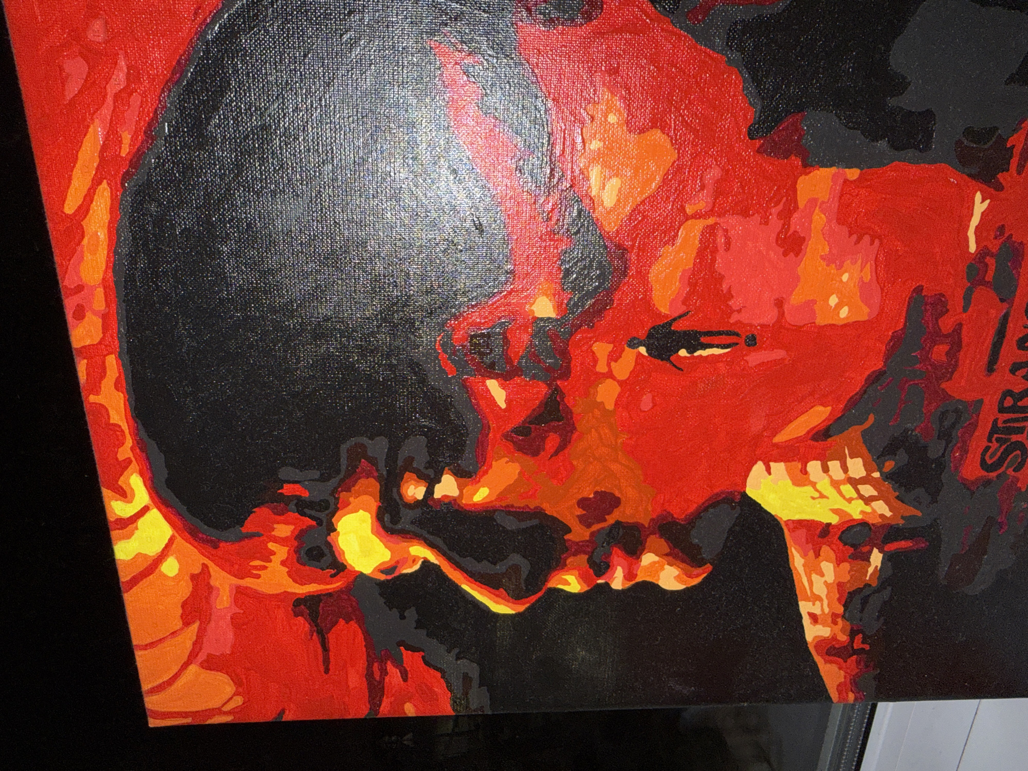 The image depicts a canvas painting with a striking color scheme primarily consisting of red, orange, yellow, and black. The subject appears to be a stylized, abstract portrait of a face or skull rendered in contrasting hues. The black areas create bold contours and shadows, while the red, orange, and yellow tones suggest warmth and intensity. The style is reminiscent of paint-by-number aesthetics. There is text "STRAN" visible along the right edge of the painting. The painting is likely photographed indoors. The image does not contain information regarding the location in Perm, Russia.