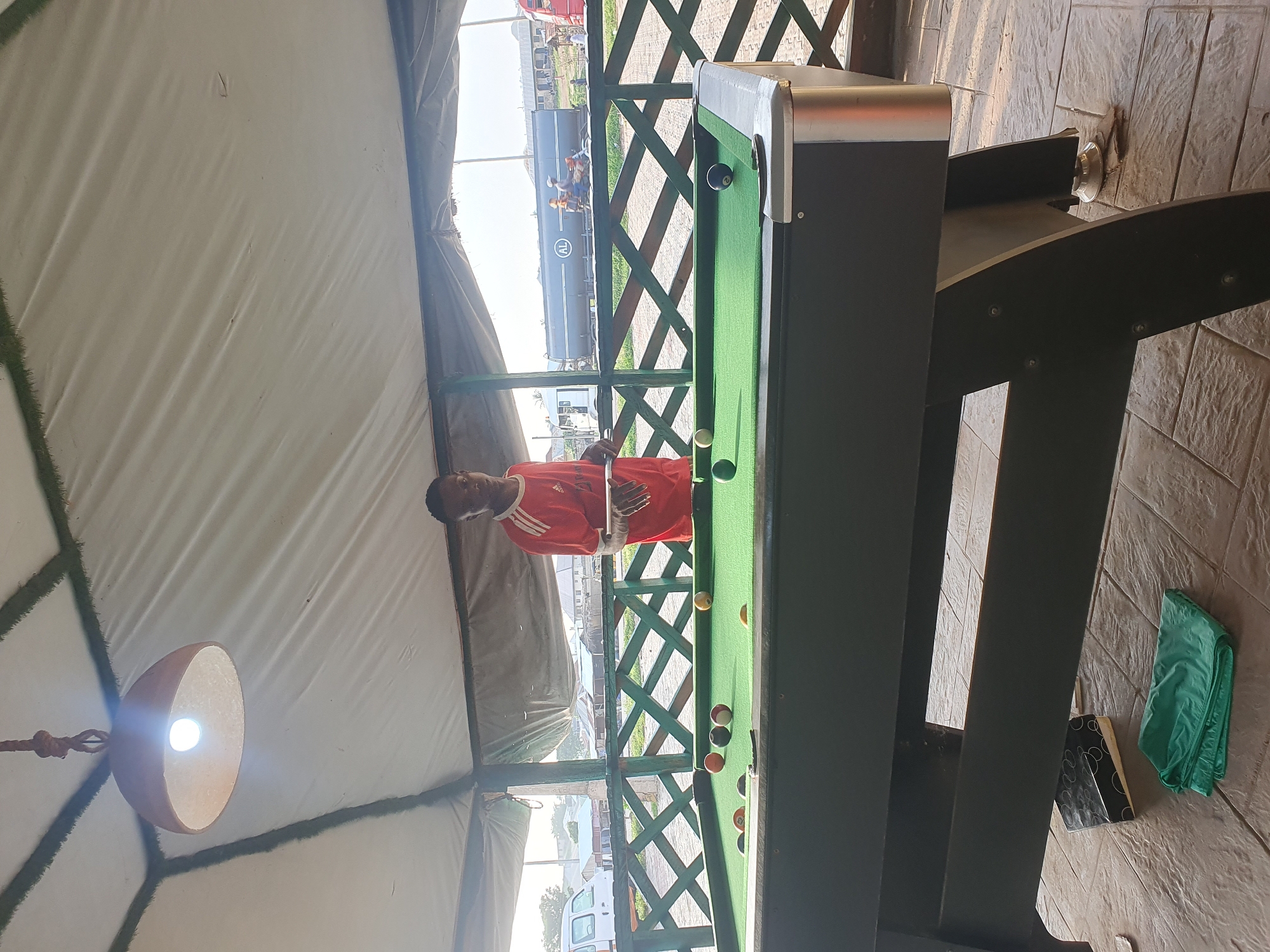 A young Black male wearing a red and white striped shirt stands behind a pool table in an outdoor covered area. The pool table is green with several balls on it, including a white cue ball. In the background, there is a wooden fence, some buildings, and a tanker truck with "AL" visible on the side. The lighting suggests it is daytime. The scene appears to be a recreational area. The presence of the specific tanker truck with the "AL" logo and the general architectural style of the buildings and fence could suggest this location is in Lokoja, Nigeria.