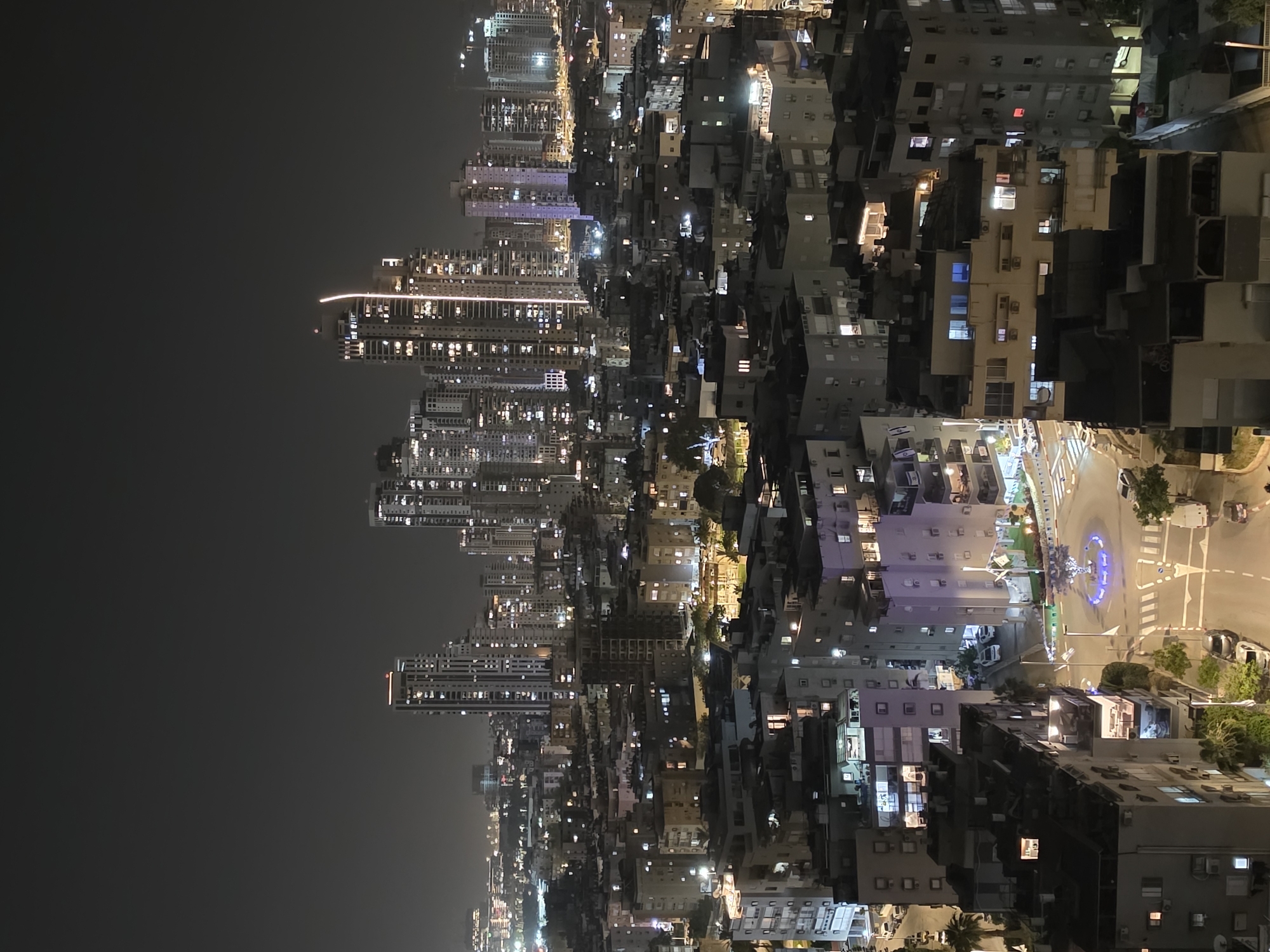 The image presents a nighttime view of Bat Yam, Israel. In the distance, illuminated high-rise buildings stand against a dark sky, reflecting light. Closer to the viewpoint, an area of predominantly low-rise buildings stretches out, their windows casting light into the night. In the immediate foreground, a brightly lit roundabout is visible, complete with vehicular traffic and surrounding greenery. The scene is lit by a mix of artificial lights from buildings and streetlights, creating a contrast between the densely built-up areas and the dark sky.