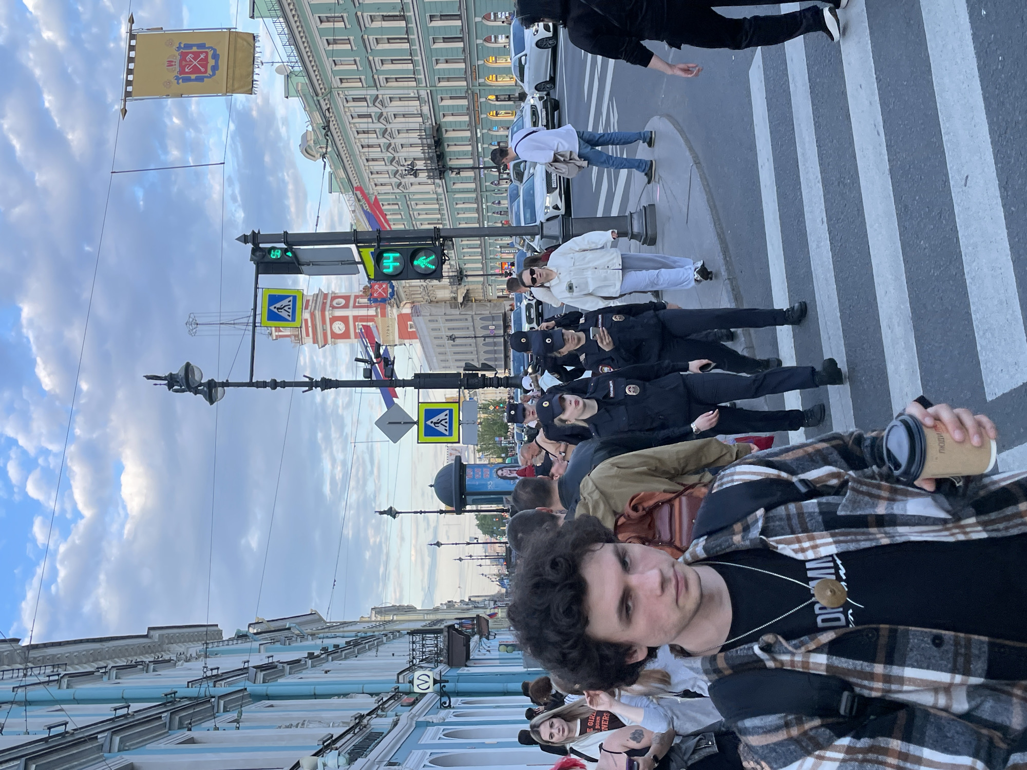 The image, rotated 90 degrees clockwise, captures a street scene in Saint Petersburg, Russia, under a cloudy sky during the day. The perspective is from street level, looking down a city street with historic architecture.
In the immediate foreground, a man with curly dark hair, wearing a plaid brown and white shirt over a black t-shirt, holds a coffee cup in his right hand. He also wears a necklace with a round pendant. Behind him and to his left, several individuals are visible, including a woman with blonde hair and a black shirt partially obscured by other people.
Further back, a group of people in dark uniforms, identified as police officers by their caps and attire with Cyrillic script (likely "Полиция" - Police), are standing or walking near a zebra crossing. One officer looks directly at the camera. Other pedestrians are also seen in various states of walking or standing on the sidewalk and crossing the street. A person in a white jacket and grey pants is crossing the street, and another person in a white shirt and jeans is further down the street. Cars are visible parked along the street and moving in the background.
Notable architectural features include multi-story buildings with intricate facades, some painted green/blue and white, lining both sides of the street. A prominent red brick building with a clock tower and the partially visible year "1703" can be seen. Street furniture includes traffic lights displaying a green pedestrian signal, a blue pedestrian crossing sign, and ornate lampposts. Overhead, a yellow banner with a crest (likely the flag of Saint Petersburg) hangs, along with smaller Russian flags or ribbons. A round blue sign with a white "M" symbol (indicating a Metro station) is also visible on a building. Text on the foreground man's t-shirt reads "DR MIN..." and another person's t-shirt partially shows "THE GRL OWNERS". Cyrillic script is also present on the coffee cup.