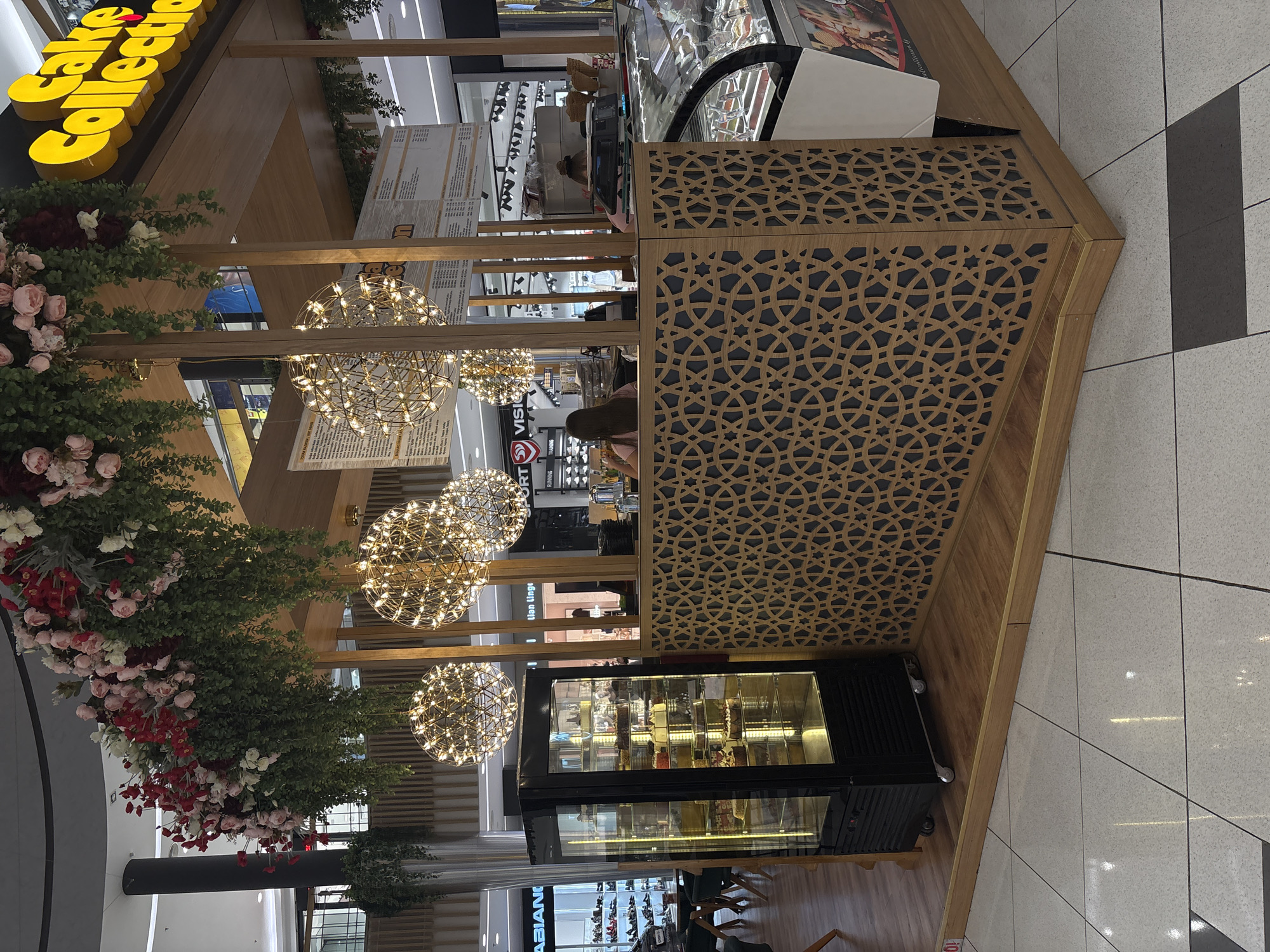 The image depicts an indoor scene of "Cake Collection," likely a kiosk or small shop in a mall or shopping center. The location is Plovdiv, Bulgaria, though this is outside the scope of what can be determined from visual elements alone.
The shop features a yellow "Cake Collection" sign on its facade. The shop's design includes wooden structures with decorative cut-out patterns. There are floral arrangements of roses and greenery around the kiosk. Three globe-shaped, modern-style chandeliers hang above. Inside the kiosk, there is a display case containing cakes and other baked goods. A person is visible behind the counter, possibly a shop attendant.
The floor is tiled in a light color with dark gray/black accents. Other stores and mall areas are visible in the background through the kiosk's structure. A customer is standing in front of the counter.
