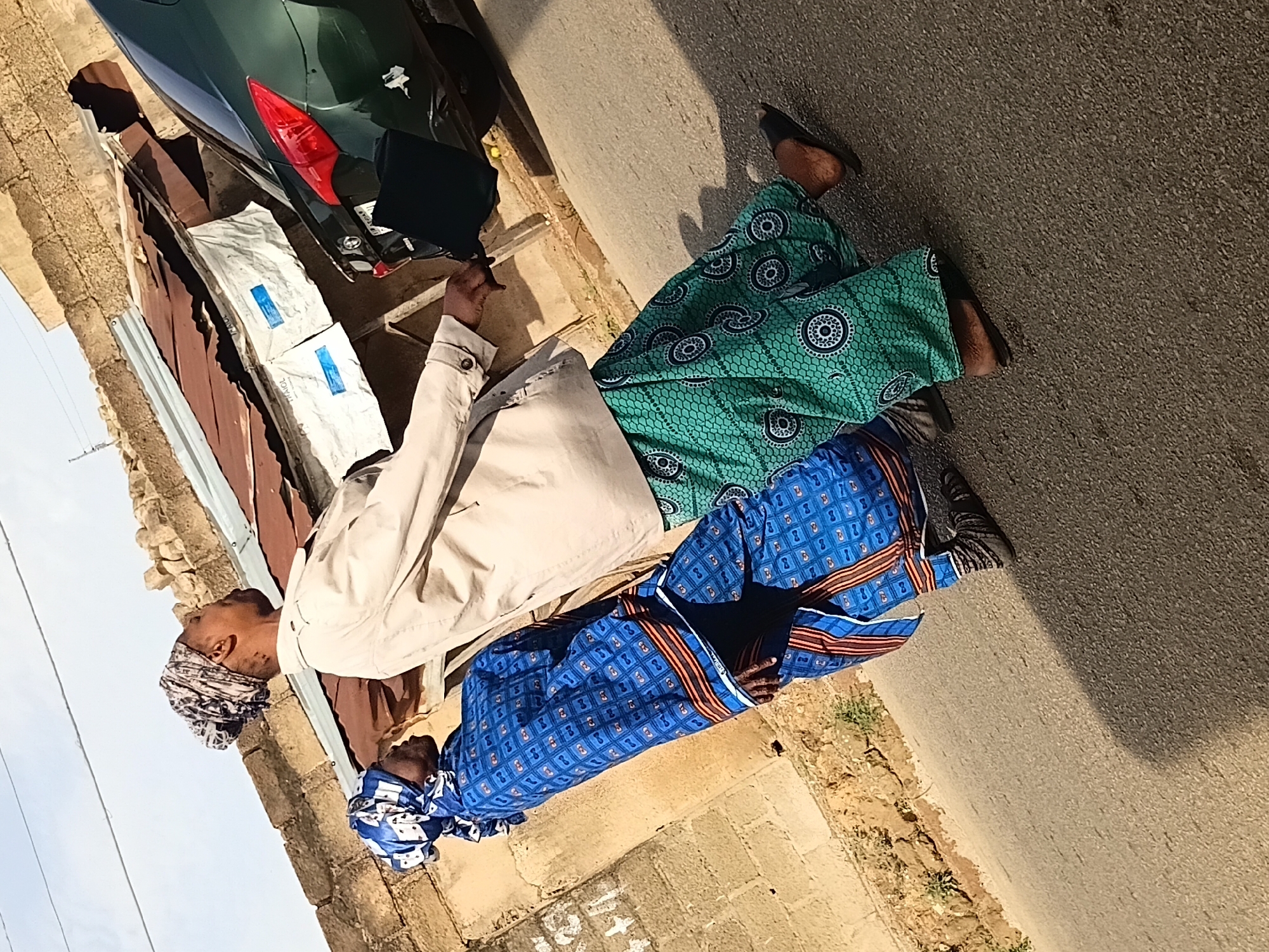 In an outdoor, urban environment, two individuals are captured in a full-body shot, walking. The person in the foreground, on the right, is wearing a light-colored jacket over a green patterned skirt and black sandals. The individual on the left is dressed in a blue patterned robe and a blue and white patterned headscarf. The background features a brick wall, a corrugated metal roof with white tarps, and a green car. The scene is illuminated by daylight, casting shadows on the asphalt ground.