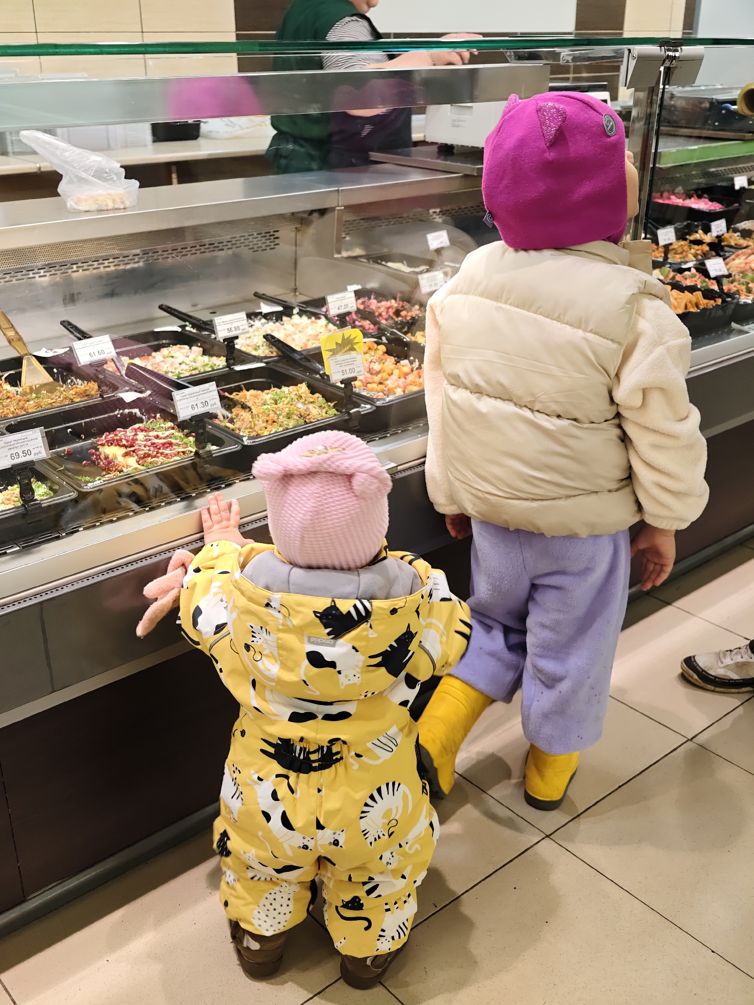 In Chaikovsky, Russia, two children are standing in front of a refrigerated display case in what appears to be a grocery store. The younger child, wearing a yellow jumpsuit patterned with cat illustrations and a pink hat, reaches toward the glass. The older child, in a cream-colored puffer vest, lavender pants, and a magenta hat, stands beside them. Both children wear yellow boots. The display case contains various prepared salads with price tags. A person in a green uniform is visible behind the display case. Another person's legs and shoes are partially visible on the right side of the image. The floor is tiled.