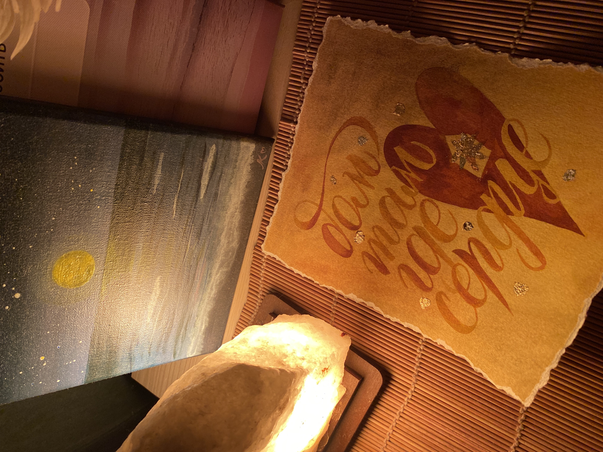 The image shows a still life composition. In the foreground, there's a rough-hewn salt lamp emitting a warm, yellowish light. Next to it is a canvas painting depicting a night sky with stars and a moon. Behind these, a handwritten card with a heart design and Cyrillic text is placed on a bamboo mat. A blurred section of fabric, possibly pink, is visible in the upper left corner. The environment appears to be indoors, with warm lighting emphasizing the textures and colors of the objects. The image was taken in the city of Chaikovsky, Russia.