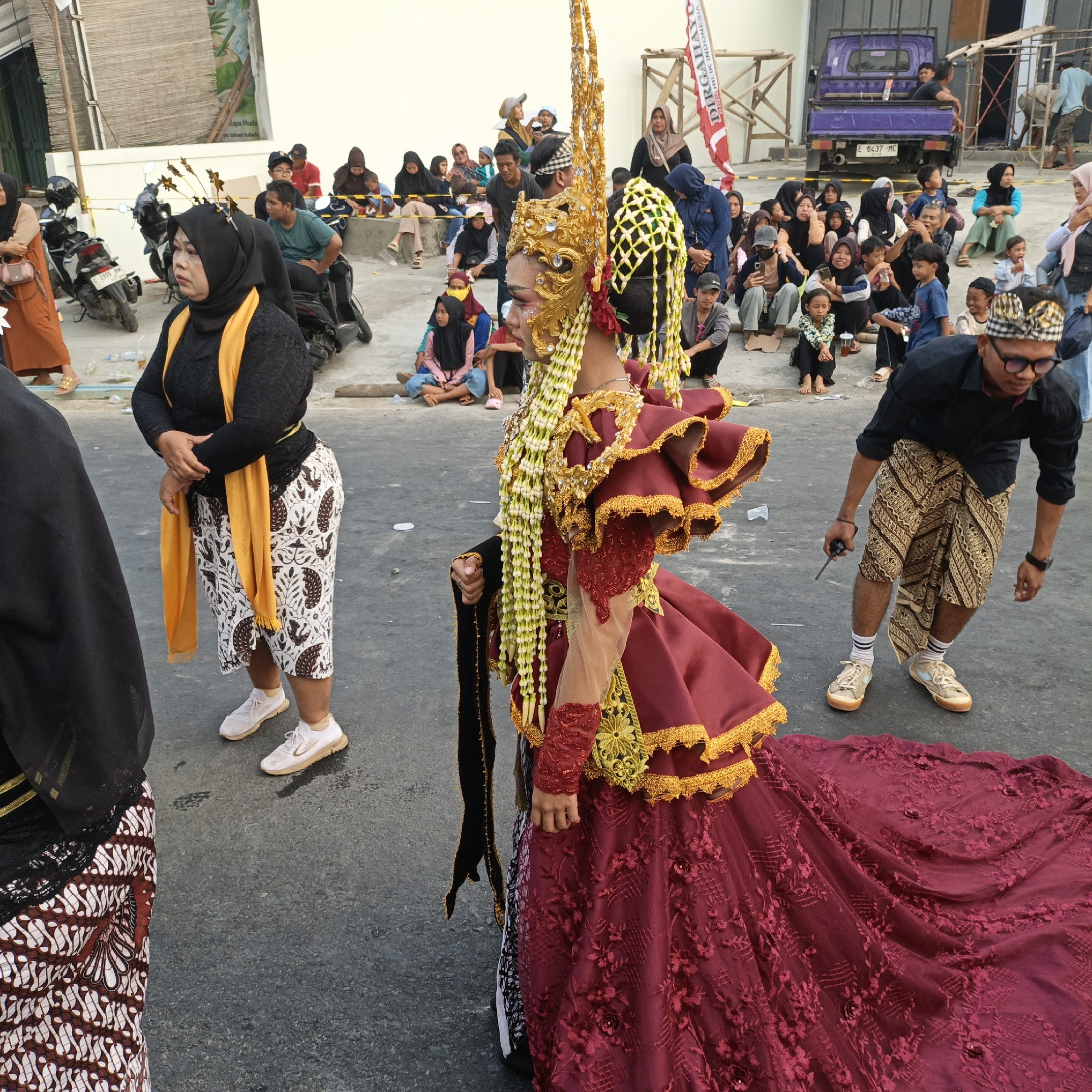 The image depicts an outdoor scene, likely in Randudongkal, Indonesia, featuring a cultural event or parade. The central focus is a person adorned in a striking, ornate costume, including a golden headdress and a flowing, deep red gown with intricate gold trim. The costume appears to be of traditional or ceremonial design. To the left, another individual is partially visible, wearing a black hijab and a yellow sash.
In the background, a crowd of spectators is seated and standing, many wearing hijabs and traditional attire, indicating a local gathering. Several motorcycles are parked in the area, suggesting a common mode of transportation. A purple truck is visible in the distance, along with scaffolding and building structures, hinting at ongoing construction or nearby infrastructure. A red and white flag with Indonesian text is also present. The ground is paved asphalt. The overall atmosphere suggests a community celebration or cultural presentation.