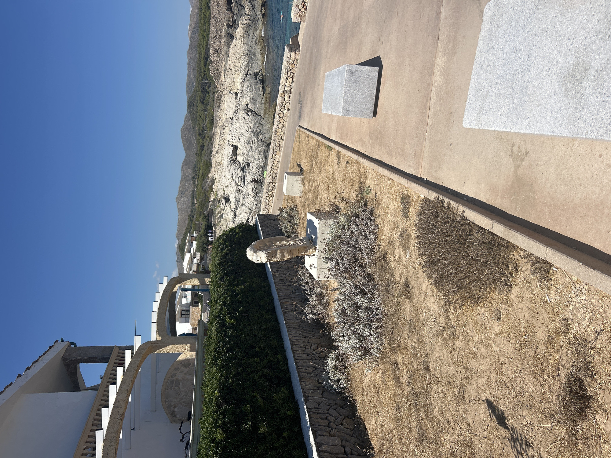 The image shows a view of the coastal area of Cala Lliteres, Spain, under a clear blue sky. In the foreground, a paved walkway runs alongside a dry, sandy area with sparse vegetation. On the walkway, there are two cubic stone benches. To the left of the walkway, a low stone wall topped with a dense green hedge separates the paved area from a patch of dry, brown grass and some withered plants. A single abstract sculpture, appearing to be made of light-colored stone, stands on a concrete pedestal in this dry area. Behind the hedge, a white building with arched balconies and stone accents is visible. Further in the background, a rocky coastline meets the turquoise sea, with a few people visible near the water and on the shore. A line of trees and hills forms the horizon.