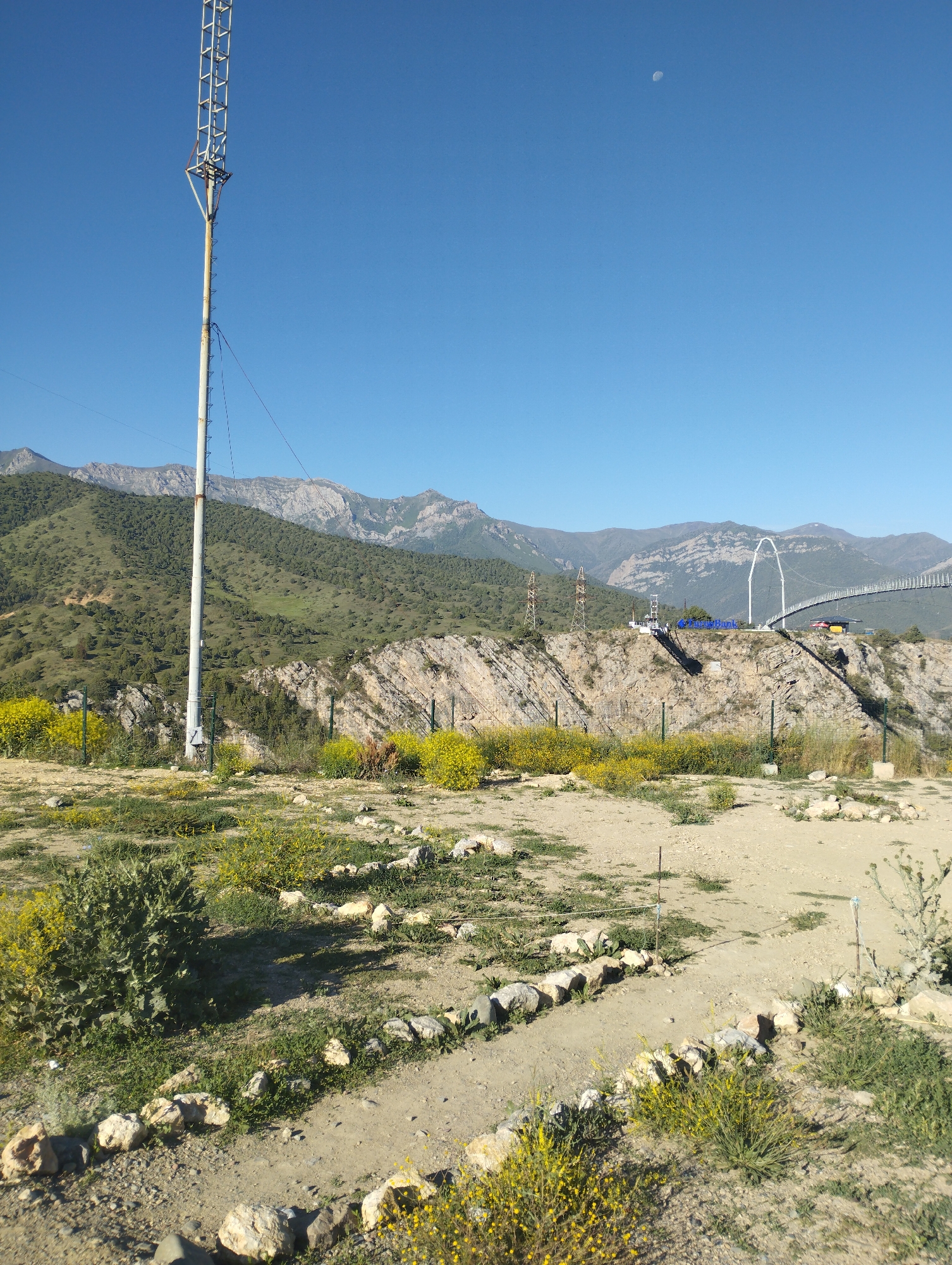 The image shows a wide, clear, blue sky with a crescent moon visible in the upper right quadrant. Dominating the left side of the frame is a tall, metal communication tower with a white pole. In the middle ground, a rocky, uneven terrain slopes upwards towards a range of green, tree-covered mountains under the bright sky. A modern, white suspension bridge with a distinctive arch is visible in the distance, spanning across a valley. The foreground is a dry, dirt and gravel path lined with rough stones, with sparse green vegetation and small yellow wildflowers scattered throughout. The environment appears to be a rural or mountainous area, possibly near a park or tourist attraction, given the bridge's design. There are no people or animals visible in the scene. The location is identified as Dardar, Uzbekistan.