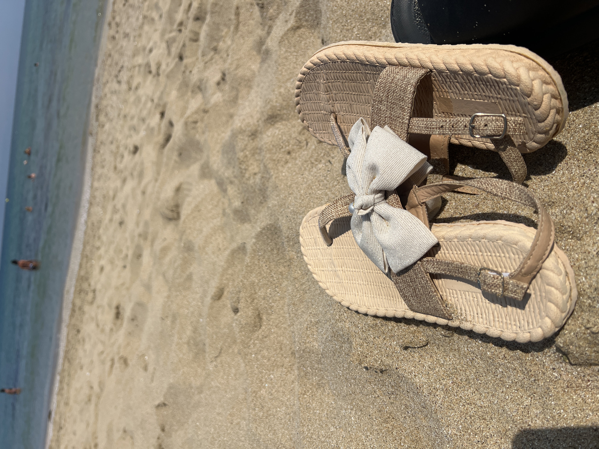 Two light brown flip-flops are positioned on a sandy beach, with the ocean and distant figures visible in the background. One flip-flop is partially obscured by the other. The flip-flops have textured footbeds and thin straps, one of which is adorned with a light-colored bow. The sand is golden and appears dry, with some footprints visible. The water is a clear blue-green. In the distance, several blurry figures are in the water, suggesting it is a warm day at the beach. The location is the city of Sozopol, Bulgaria.