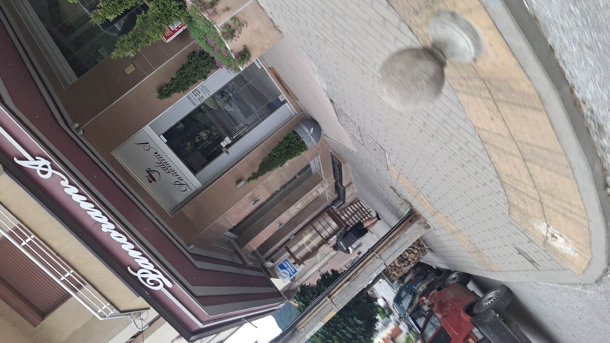 The image shows the exterior of a cafe named "Panorama A" located in Gotse Delchev, Bulgaria. The cafe has a prominent sign with the name "Panorama A" in white cursive lettering. The entrance to the cafe is a glass door with "Cafe Panorama A" written on it, along with what appears to be operating hours. There are two small green conical trees planted in large grey pots outside the cafe. A red car and a dark colored car are parked on the street in front of the cafe. A person wearing a black jacket and shorts is walking on the sidewalk away from the cafe. The sidewalk is paved with interlocking bricks and has a yellow-colored border. A concrete sphere on a pedestal is visible on the sidewalk. The building facade is made of beige-colored tiles. The overall scene depicts a street view in front of a business.