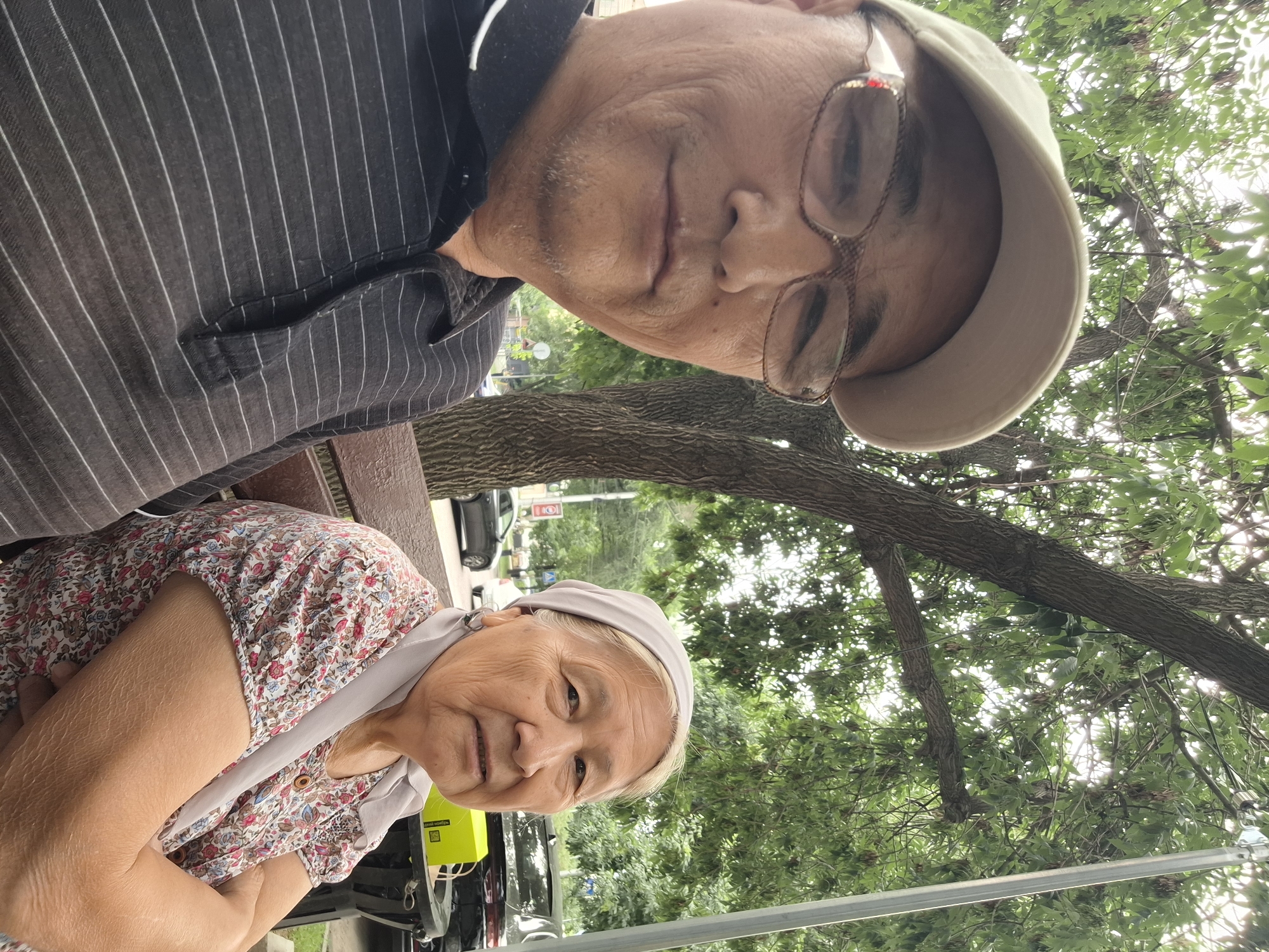 A close-up, selfie-style shot shows two individuals, likely an older couple, seated outdoors on a bench. The person in the foreground on the left, wearing a beige baseball cap and glasses, is looking at the camera. They are wearing a dark, pinstriped polo shirt. To the right, an older woman with a light-colored headscarf and a floral print blouse is also looking towards the camera, with her arms crossed. They are positioned beneath a tree with lush green leaves, and in the background, there are cars and a street visible, suggesting an urban setting in Almaty, Kazakhstan. The lighting indicates it is daytime.