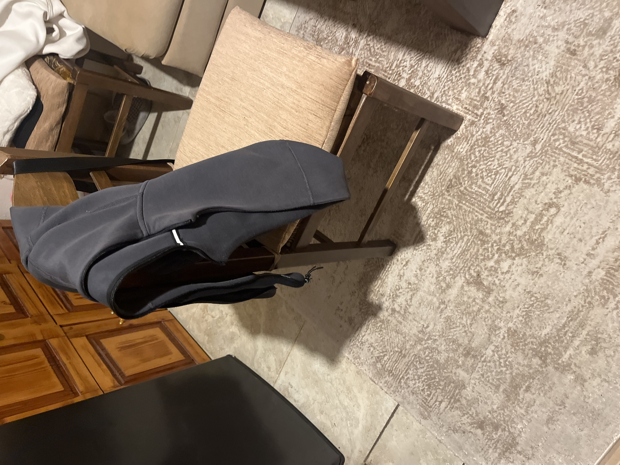The scene depicts an interior room, likely a common living space, observed from a slightly elevated, oblique perspective. A dark grey hooded jacket with a front zipper and a white accent on the zipper pull is draped over the back of a wooden chair situated centrally in the mid-ground. This chair features a light brown, upholstered seat cushion. To the left, a large, dark, rectilinear object, possibly a refrigerator or cabinet, partially extends into the frame. In the background to the left, a reddish-brown wooden cabinet with multiple paneled doors and small golden knobs is visible. To the right of the central chair, a second, similar wooden chair is present, stacked with light-colored, soft items or cushions. Partially visible beneath this second chair is a light-colored shoe. Further to the right, a section of a light brown upholstered sofa or armchair is present. The floor consists of light-toned, possibly beige or cream, tiled sections with visible grout lines. A rectangular area rug with a textured, abstract pattern in similar light brown and cream tones covers a portion of the tiled floor in the foreground. Artificial lighting casts distinct shadows from the furniture onto the floor and rug. No individuals are present in the image. The scene is an interior room within the city of Blagoevgrad, Bulgaria.