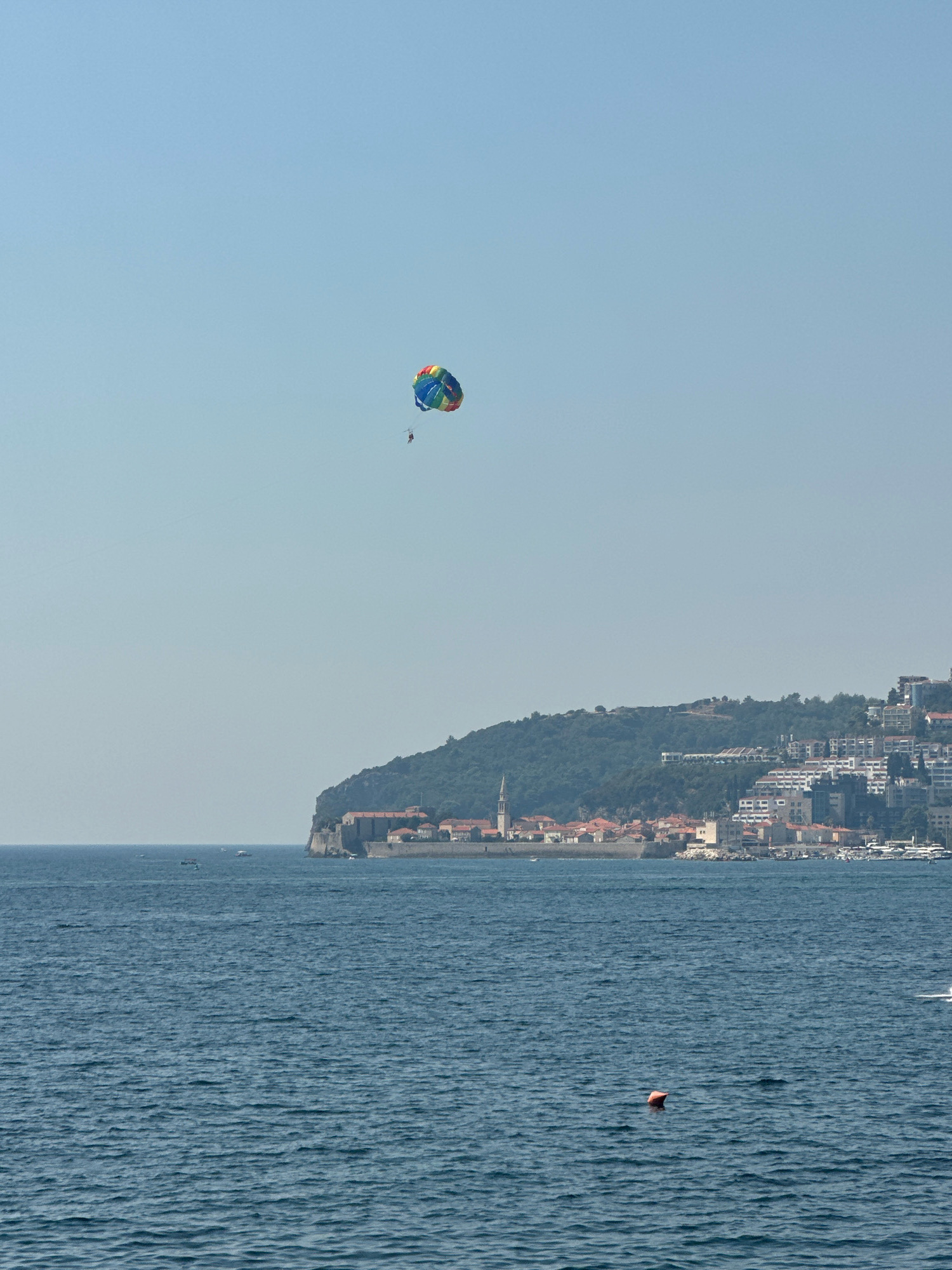 The image depicts a coastal scene in Budva, Montenegro. In the clear blue sky, a colorful parasail with a person attached is visible, appearing to be engaged in a recreational activity. Below the parasail, the turquoise waters of the Adriatic Sea stretch towards a hilly coastline. On the coastline, the historic walled Old Town of Budva is situated, characterized by its red-tiled roofs and stone buildings. Adjacent to the Old Town, modern white buildings ascend the hillside, indicating urban development. Several small boats are scattered across the water. A small orange buoy is floating on the sea's surface. The overall impression is a bright, sunny day with a blend of historical and modern architecture along the picturesque Montenegrin coast.