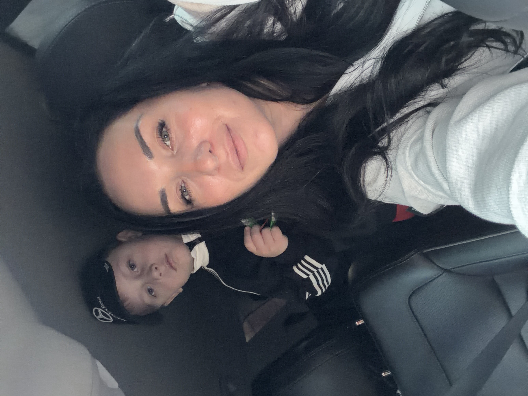 The image captures an adult female and a young child in a selfie taken inside a car. The adult, positioned in the upper right and appearing to be taking the photo, has long dark hair and is wearing a light-colored, possibly white, hooded top. Her head is tilted, and the image is rotated, showing her looking towards the camera with a subtle smile.
To her lower left, a young child is visible, also looking towards the camera with a neutral expression. The child is wearing a black beanie that prominently displays a white Mercedes-Benz logo and the text "Mercedes-Benz". They are dressed in a black top with white stripes on the sleeve, characteristic of sportswear. The child holds a small, indistinct object with green and red elements in their right hand.
The setting is the interior of a vehicle, identifiable by black leather car seats and a visible seatbelt. The lighting suggests it is daytime, though no specific outdoor details or weather conditions are discernible. The image conveys a casual, intimate moment. The provided location context for this media is Dzerzhinsk, Russia.