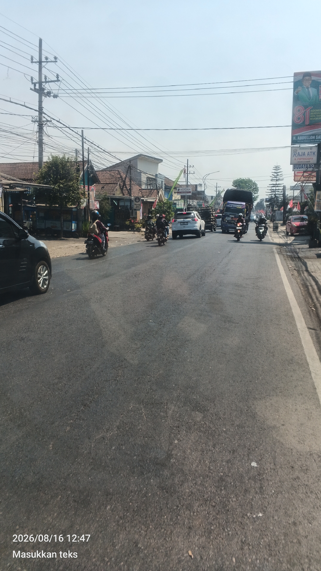 The image depicts a street scene in Bunut Wetan, Indonesia, on a clear, sunny day. The foreground is dominated by an asphalt road with a white line marking the edge on the right. Several vehicles are present, including a black car on the left, a white SUV, a dark truck, a red car, and multiple motorcycles. Several individuals are riding the motorcycles. On the left side of the road, there are buildings with red tile roofs, trees, and utility poles with numerous wires. On the right side of the road, a large billboard displays an image of a man and text. The overall atmosphere suggests a typical day in a moderately busy urban or semi-urban area.