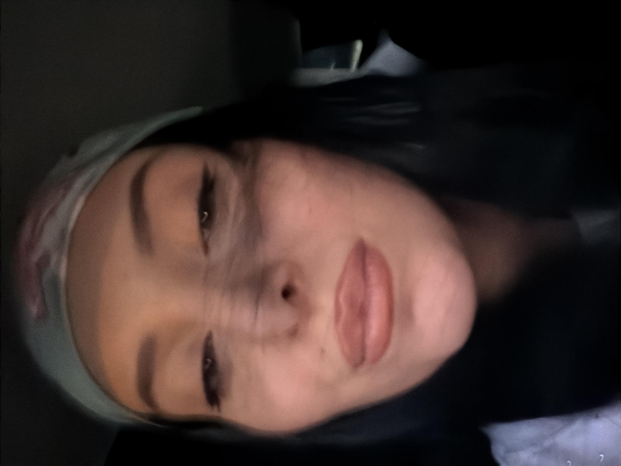 A close-up, slightly blurred, indoor selfie of a person with dark hair and fair skin. They are wearing a light-colored bandana with a subtle pattern. Their eyes are dark, with visible eyeliner. Their lips are full and painted a soft pink. The background is dark, suggesting an evening or dimly lit setting. No specific location in Tbilisi, Georgia, can be identified from the image.