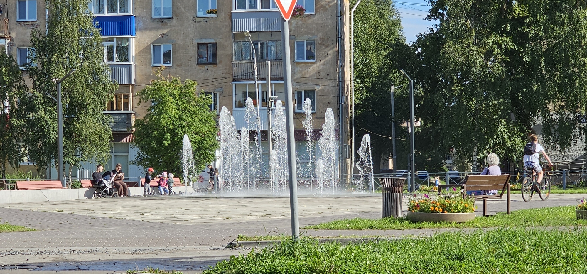 The image depicts a sunny day in a city park in Novodvinsk, Russia. In the foreground, a paved area features a multi-jet fountain with water spraying upwards. To the left of the fountain, several children in casual attire are playing near a red bench. A family, consisting of an adult male and female, is seated on another red bench, with a stroller beside them. In the background, a multi-story apartment building with a weathered facade and balconies is visible, partially obscured by trees. Further to the right, a woman with grey curly hair is seated on a wooden bench, facing away from the camera. A person is riding a bicycle on a path behind her, also facing away. Flowerbeds with orange and pink flowers are present in the foreground and midground. A trash receptacle stands near the fountain. The overall scene suggests a recreational area within a residential urban environment.