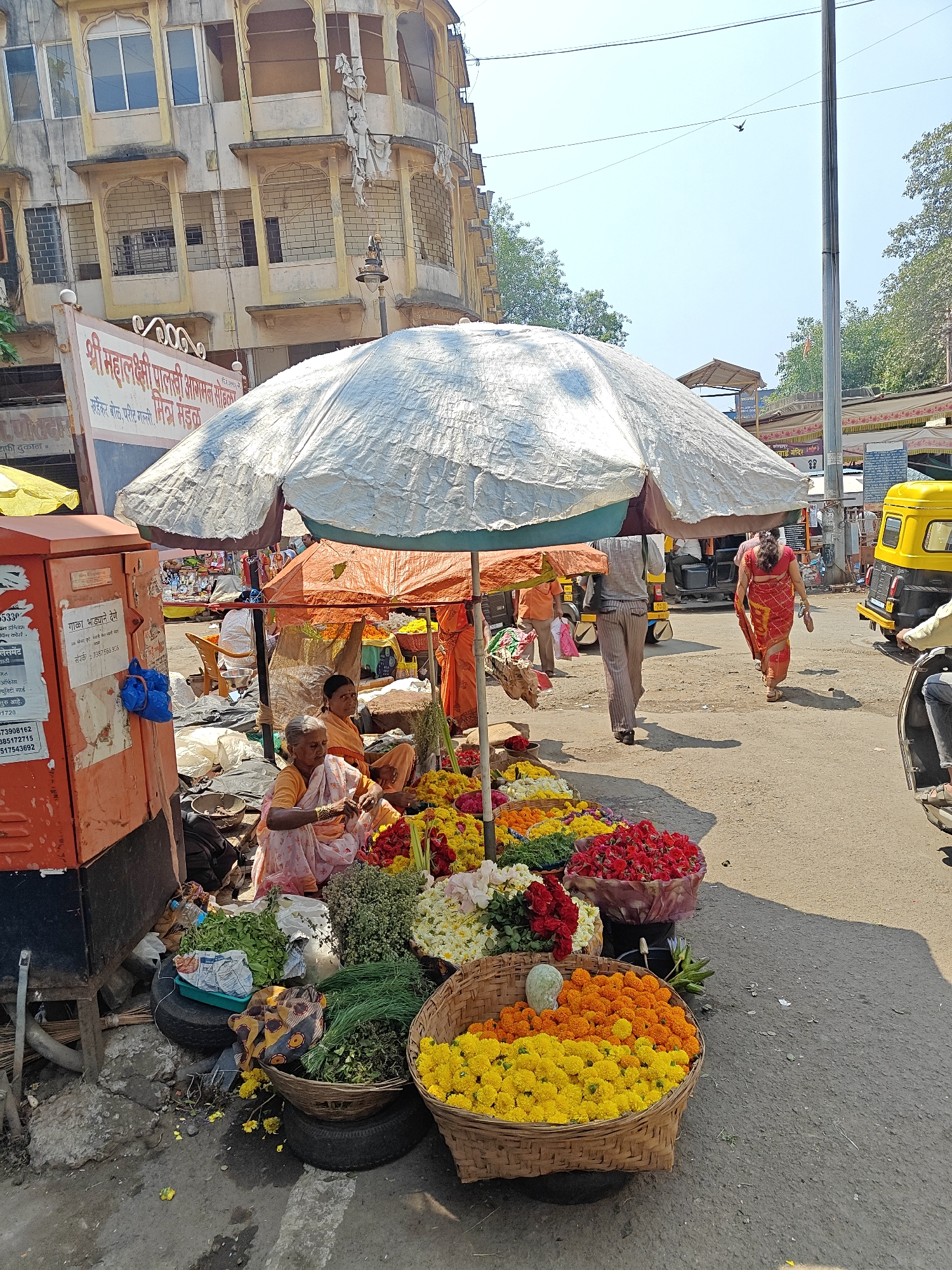This image depicts a bustling daytime street market scene in the city of Unknown, Unknown, specifically identified by a sign as potentially being on Vader Bol, Parit Galli, near a Shri Mahalakshmi Palkhi Aagaman Rangoli. The ground consists of a dry, unpaved asphalt or concrete surface with scattered debris and flower petals. Overhead, the sky is clear and bright blue, with a lone bird in flight visible.
In the foreground and midground, a flower market operates under large canvas umbrellas, including a prominent white tarp umbrella and smaller orange ones. Two elderly women are seated on the ground under the main white umbrella. One woman, wearing a pink and white patterned sari with an orange blouse, is positioned in the immediate foreground, actively engaged with sorting or arranging flowers. Behind her, another woman in an orange sari is seated. Numerous woven baskets containing a variety of fresh flowers are arranged around them and extend towards the right. These include large quantities of yellow and orange marigolds, red roses, white flowers, and baskets of green herbs or foliage. Used automobile tires serve as makeshift stands for some baskets. To the far left, a tall, orange metal utility box or post box is visible, adorned with taped notices in Marathi and a blue plastic bag hanging from it.
Pedestrians are observable on the street. One individual, dressed in a light-colored shirt and striped trousers, walks away from the camera towards the background. Further right, a woman in a red sari walks away, holding a pair of sandals in her left hand. Several yellow and black auto-rickshaws are present on the street, one partially visible on the far right. In the background, a multi-story building with a light-colored, worn facade features multiple windows and decorative arched balconies. Dense green trees are visible in the background, along with additional market activity and structures. A tall utility pole stands on the right side of the frame, with overhead power lines. Shadows are cast strongly by the market umbrellas and individuals, indicating direct sunlight.