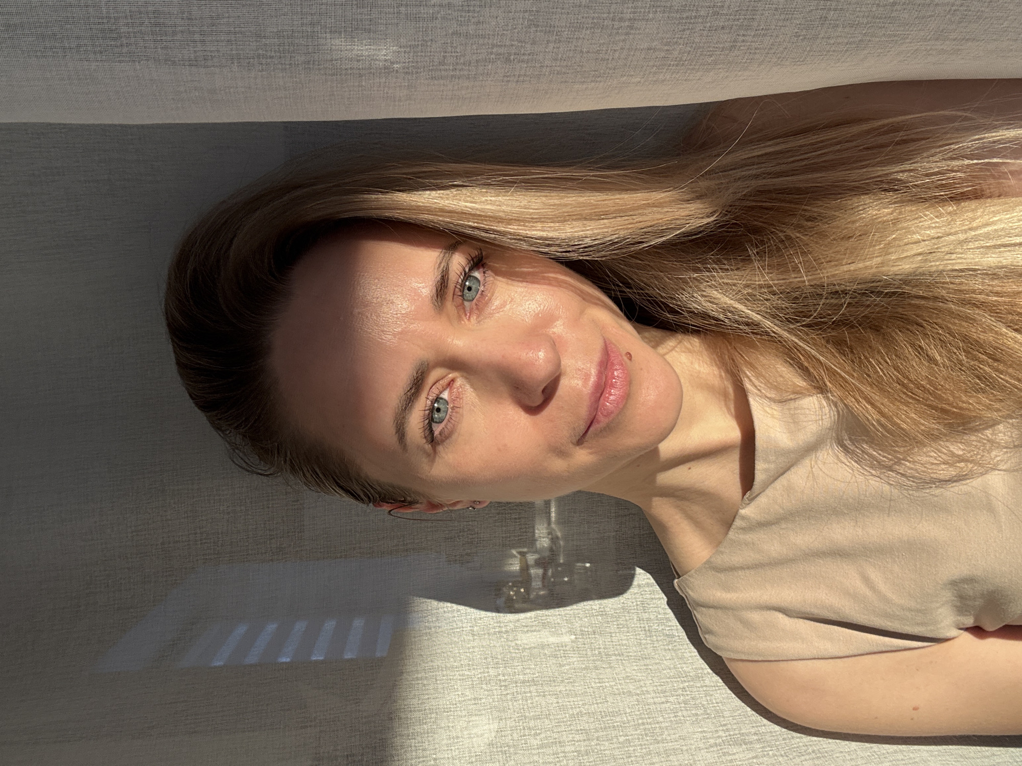 A light-skinned woman with blond hair and blue eyes lies on a textured, light gray surface. Her hair is spread out around her head. She is wearing a beige top. Light from a window casts shadows on the surface behind her head, creating a striped pattern. The background appears to be draped with a sheer fabric. No specific details in the photo confirm the location as the city of Chaikovsky, Russia.