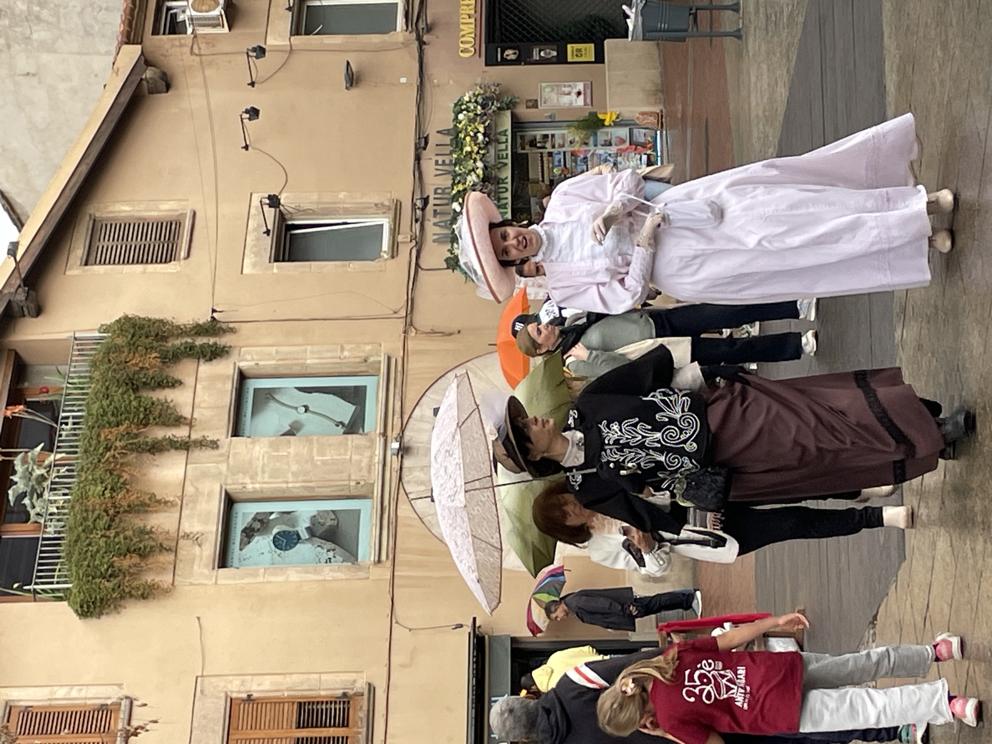 The image captures a group of people on a paved street in front of a building. One woman, wearing a large pink hat and a long white dress with pink trim, stands prominently. Another woman stands behind her in a long brown skirt and dark jacket with white swirls. Others are present, some holding umbrellas. The building behind them has windows with shutters and window boxes with flowers and plants. There's a sign on the building that reads "NATUR VELLA". Based on this sign and the architectural style of the building, it is likely in Terrassa, Spain, near the Natur Vella shop. The people in the image appear to be dressed in vintage clothing, possibly for a historical event or performance.