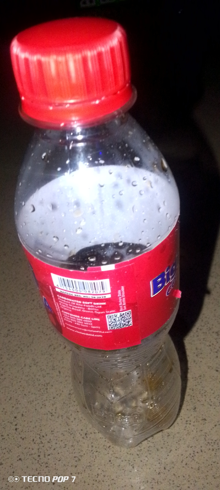 A red-capped plastic bottle of carbonated soft drink sits on a speckled countertop in an indoor setting. The bottle is partially filled with a dark liquid and condensation is visible on its exterior. A red label with white lettering and a barcode is wrapped around the upper portion of the bottle, partially obscured by water droplets. The bottle appears to be upright. The background is dark and out of focus, with a shadow cast from the bottle. The bottom left of the image displays a "TECNO POP 7" watermark.