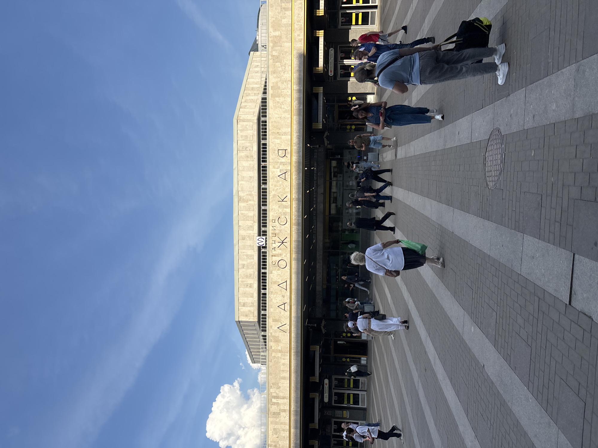 This image displays the exterior of Ladozhskaya (Ладожская) Metro and Railway Station in Saint Petersburg, Russia, under clear daytime conditions.
The prominent central structure is a multi-story building clad in light-colored, possibly beige or cream, stone panels. The station's name, "ЛАДОЖСКАЯ," is spelled out in large, dark Cyrillic letters affixed to a horizontal band on the main facade above the entrance level. A circular blue logo with the letter "M," signifying a metro station, is visible to the right of the text on the upper facade. The ground level features multiple dark-framed glass entrances and exits, some with yellow and black striped markings indicating designated pathways.
In the foreground, a large paved plaza extends from the station entrance. The paving consists of alternating wide stripes of light gray and darker gray (or possibly reddish-brown) rectangular pavers. A circular metal manhole cover is visible in the lower-middle section of the plaza.
Numerous pedestrians are observed on the plaza, moving in various directions, primarily entering and exiting the station. Individuals are dressed in casual attire. Notable figures include: a woman in a white top and dark skirt carrying a green bag, walking away from the station on the right; a woman in a light blue t-shirt and plaid pants, carrying a black and yellow duffel bag, also walking away from the station on the right-middle; a woman in a denim jumpsuit holding a phone, walking towards the station in the center-right; and a woman in a white two-piece outfit (crop top and wide-leg pants) walking left in the center-left. Other individuals are seen in various stances, walking or standing.
The sky above is bright blue with scattered white cumulus clouds, notably a large, fluffy cloud mass visible in the upper-left quadrant. The scene is well-lit by direct sunlight, casting visible shadows on the ground and structures.