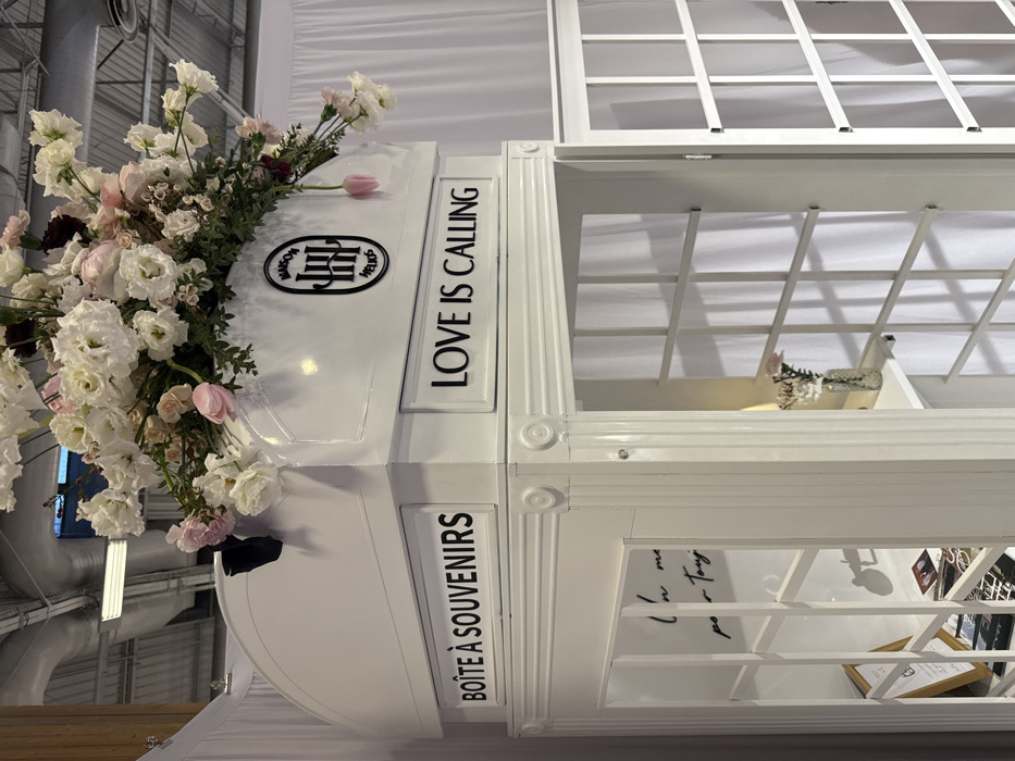 An upside-down, white, multi-tiered, enclosed structure adorned with flowers is shown. Text reading "BOÎTE À SOUVENIRS," "LOVE IS CALLING," and a circular logo are visible on the top layer. The structure features window-like bars, with objects placed inside, including a white bird. The interior also contains written text and picture frames. The surrounding background includes building structures and ceiling fixtures.