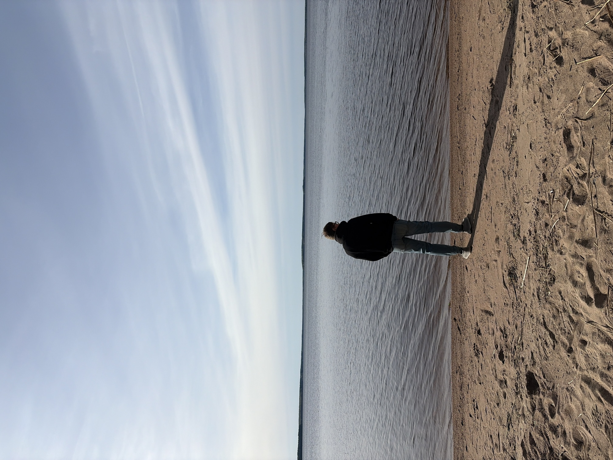 A medium-shot, vertically-oriented photograph shows a person standing on a sandy beach, looking out over a body of water. The beach takes up about one-third of the right side of the image, while the water occupies the central third, and the sky fills the left third. The person, facing away from the camera, is wearing a dark jacket and light-colored jeans. The sky is pale blue with streaks of white clouds. The water appears calm, with subtle ripples. The location is likely near the city of Glubokoe, Russia, given the context. The sand is tan and textured, with dry grass blades scattered around. A long shadow is cast by the person onto the sand.