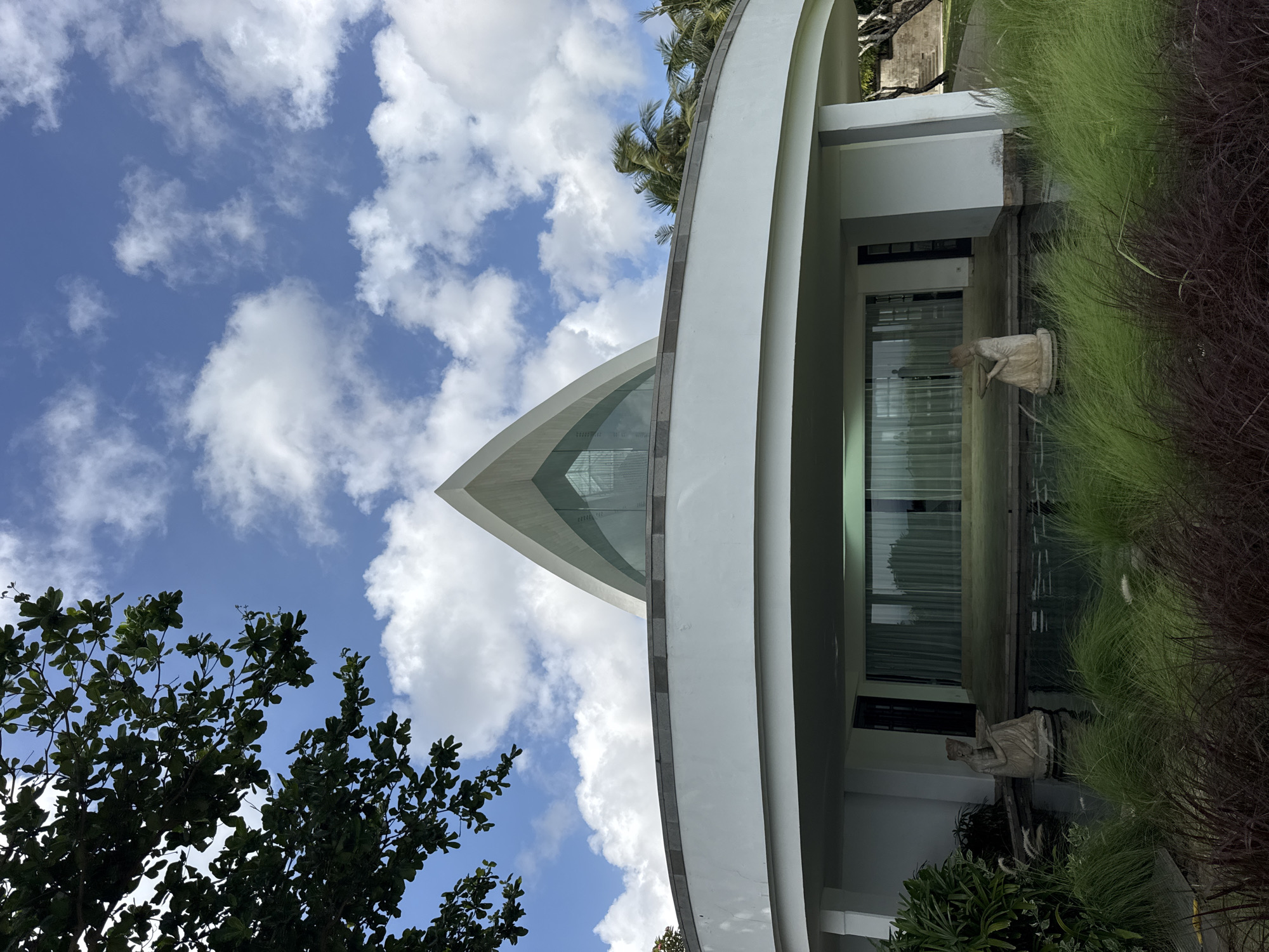 The image shows a modern architectural structure in Kuta Selatan, Indonesia. The building is characterized by a curved, white facade and a prominent triangular glass section at the top. Below the glass apex, the building features an open, covered area with glass windows. Adjacent to the building is a reflecting pool with statues and ornamental grasses. To the side, foliage from trees and other plants is visible, contrasting with the clear blue sky dotted with cumulus clouds.
