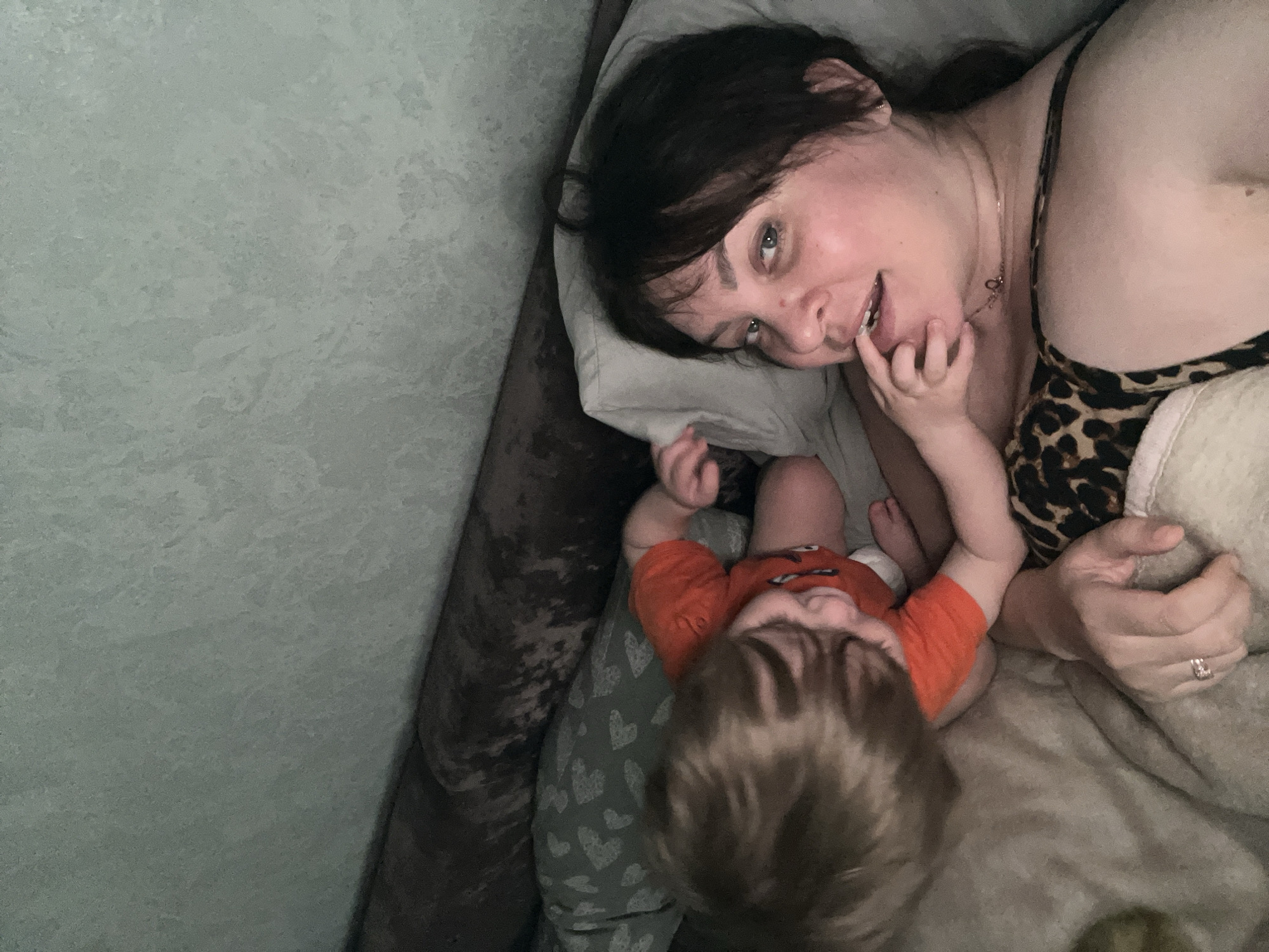 A woman and a baby are lying down. The woman is on a pillow and has her finger in her mouth, she is wearing a leopard print top. The baby is wearing an orange shirt with a cartoon on it and gray pants with white hearts. The location cannot be identified based on the image.