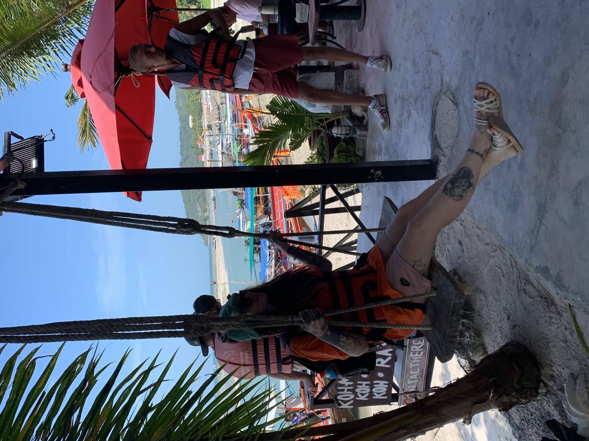 In the city of Ko Samui District, Thailand, a woman with long dark hair, wearing sunglasses, a green hat, an orange t-shirt over a life vest, and pink shorts, is sitting on a wooden swing. Her legs are crossed, and she is wearing sandals. She has tattoos on her arms and legs. Another person, a man with blond hair wearing a white shirt and a red life vest, stands to her right. Behind them, a beach is visible with numerous long-tail boats docked in the turquoise water. Palm trees frame the scene, and a red umbrella provides shade for an outdoor seating area.