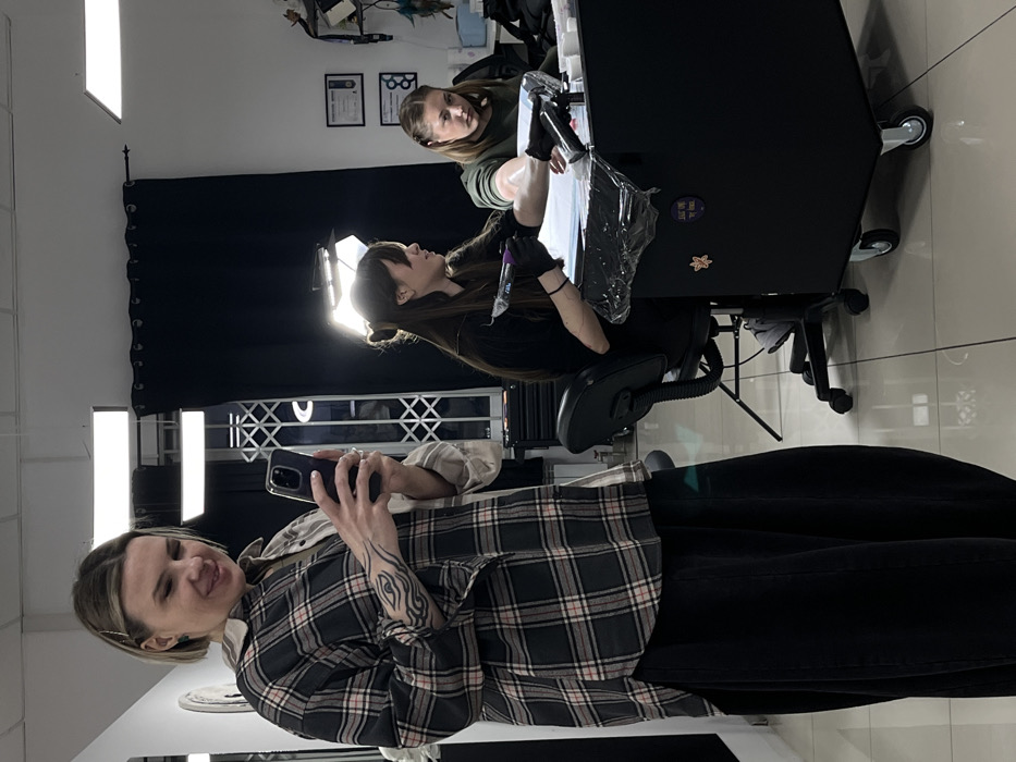 An indoor scene within a tattoo parlor in Rostov-on-Don, Russia, depicts three women. In the foreground on the left, a blonde woman wearing a plaid shirt and dark pants holds a smartphone in both hands, angled towards the right, appearing to record or photograph. She has a distinct eye-shaped tattoo on her left forearm and a slight smile. To her right, a dark-haired woman, identified as a tattoo artist, is seated in a black rolling chair, wearing black gloves and a dark top. She is actively performing a tattoo procedure on the left forearm of another woman, who is seated to her right, wearing an olive-green top. The client's arm rests on a black mobile workstation, which is covered with clear protective plastic. The workstation also holds various tattoo supplies. A large softbox light illuminates the tattooing area from behind the artist. The background features white walls, horizontal rectangular ceiling lights, a black curtain, a white lattice-like structure, and framed items on the wall. The floor is composed of reflective light-colored tiles.