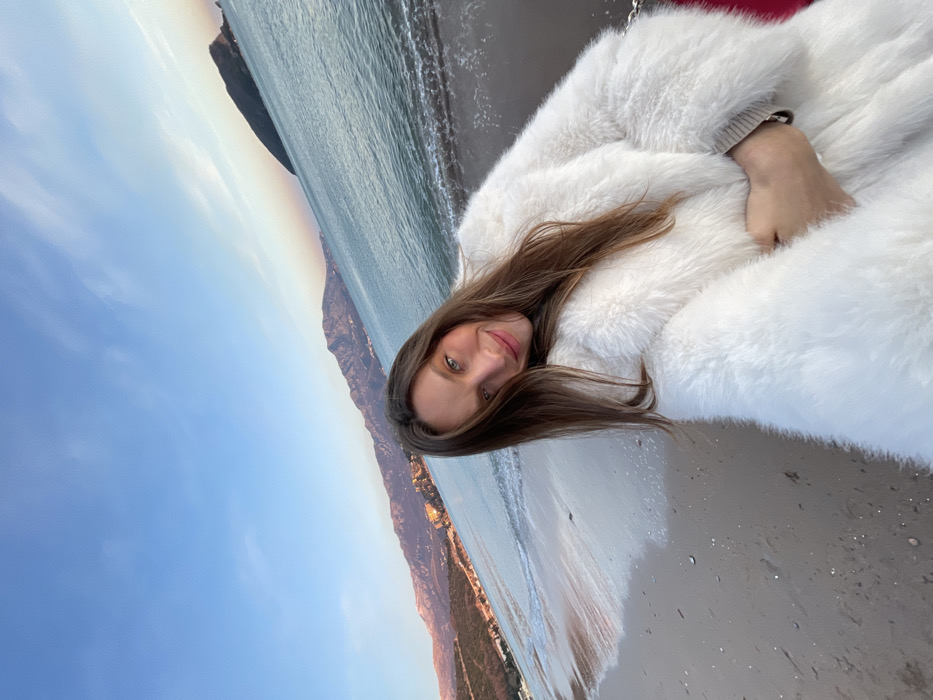 A woman with long brown hair is lying on a sandy beach, wearing a large white fur coat. The ocean stretches out behind her, with waves lapping at the shore. A mountainous coastline is visible along the horizon under a blue sky. The image is rotated slightly, giving a tilted perspective of the scene.