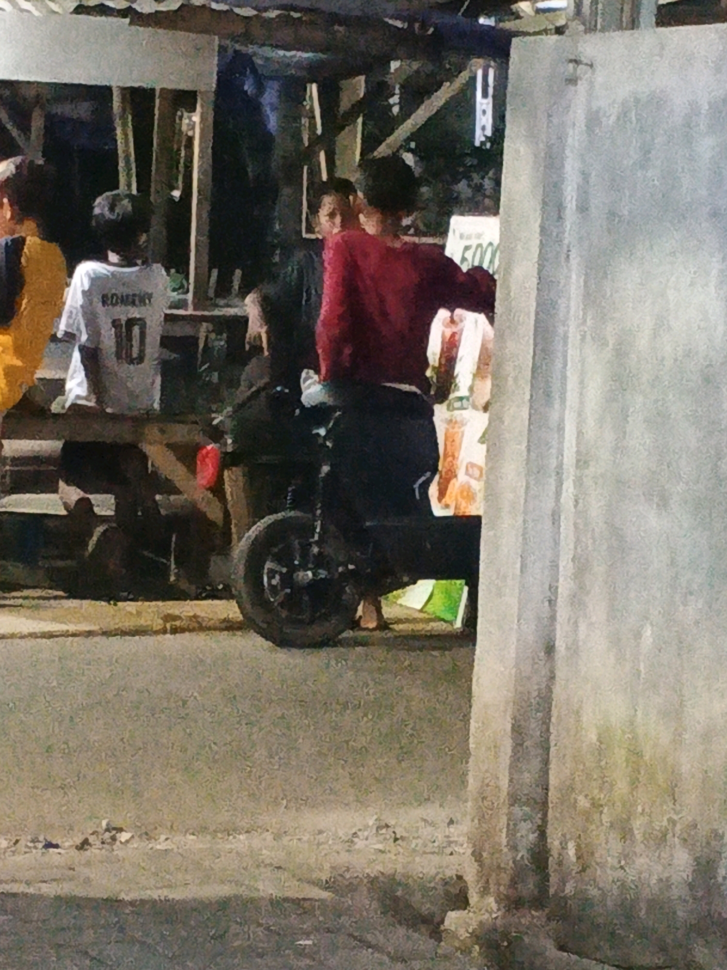 The image depicts a street scene in Kadu, Indonesia, likely at night or dusk due to the low light conditions. In the foreground, a person is seated on a black motorcycle. Behind the motorcycle, a wooden structure is visible, possibly a stall or shop, with a white sign displaying some text and numbers, suggesting a commercial area. Several individuals are present, some standing and others seated. One person is wearing a white shirt with the number "10" and the name "ROMERY" on the back. The environment appears to be an outdoor, possibly urban or semi-urban setting, with a paved surface in the foreground.