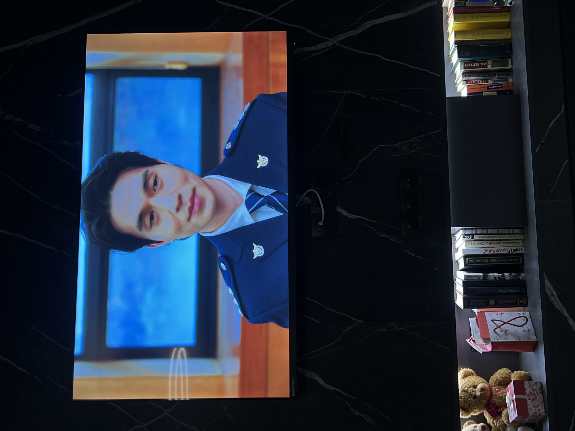 The image shows a television screen displaying a man in a blue uniform with a window behind him. To the right of the television screen, there is a built-in shelf containing a stack of books at the top, followed by more books, gift bags, and teddy bears at the bottom. The surrounding area has a dark, marbled appearance.