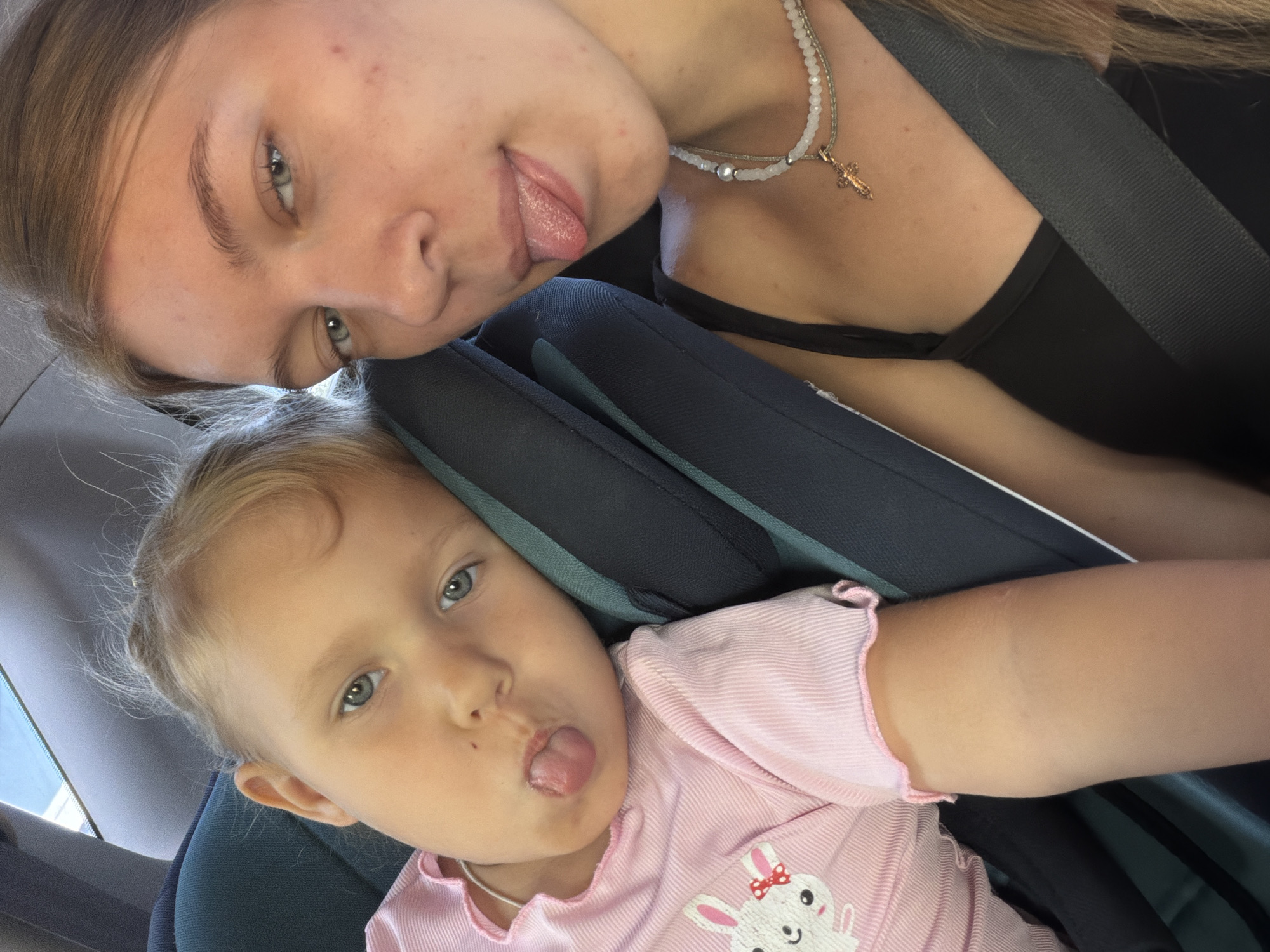 Two females are in a car. A younger female, likely a child, is seated in a car seat and is wearing a pink shirt with a rabbit graphic. She is sticking her tongue out. An older female, likely a teenager or young adult, is seated next to her, also in the car, and is taking a selfie. She is wearing a black tank top and a necklace. The car appears to be in motion, as indicated by the blurred background outside the window. The interior of the car is visible, including car seats and seatbelts. The lighting suggests it is daytime. The location of Rostov-on-Don, Russia is not depicted or implied in this image.