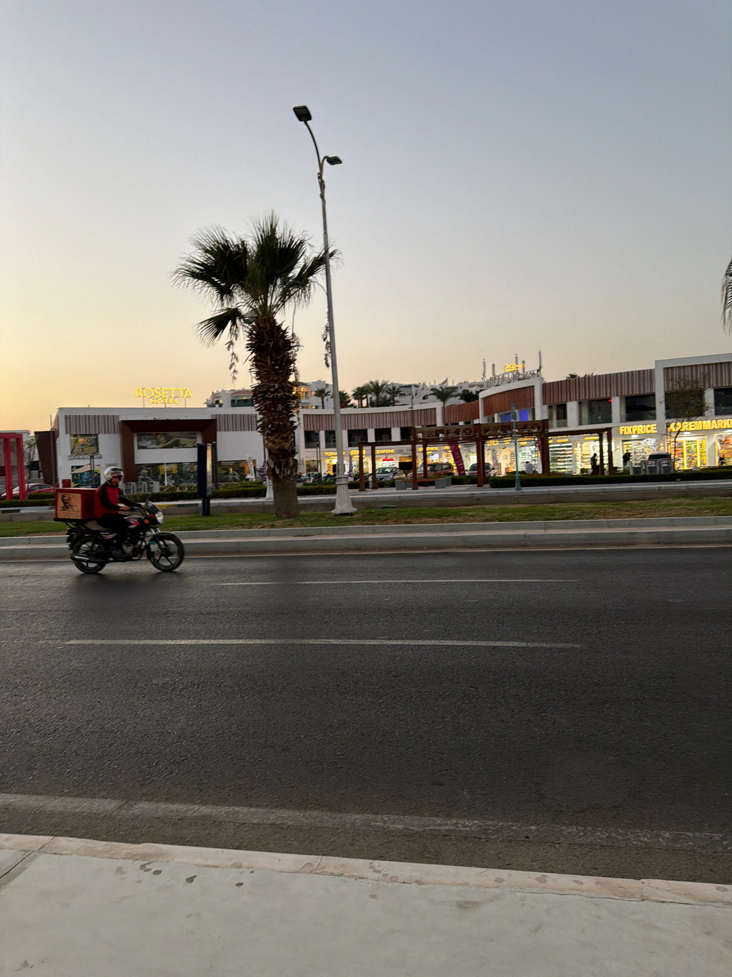 This is a medium-shot, eye-level, outdoor, vertical photograph taken during the day in Sharm El Sheikh, Egypt. The scene depicts a street in front of a commercial building complex. A man is riding a motorcycle on the asphalt road, carrying a large red box on the back. The man is wearing a red shirt and a helmet. The commercial buildings feature a sign that reads "ROSETTA HOTEL" in prominent yellow lettering. Other visible signs include "FIXPRICE" and "KOREANMARKET". Palm trees are visible in front of and around the buildings. The sky is clear and a pale blue. The road has white lane markings. A sidewalk runs along the side of the road.