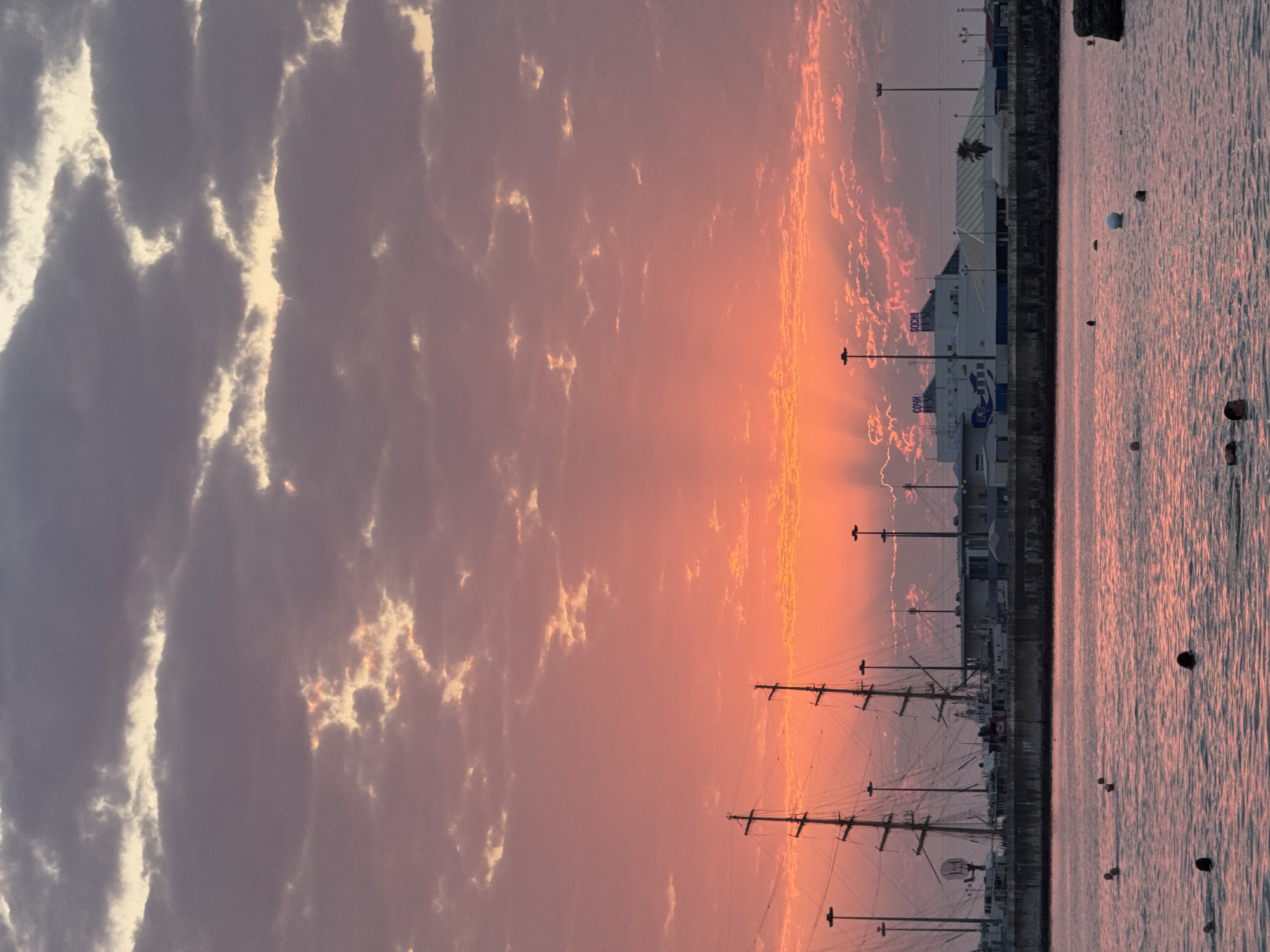 A twilight scene unfolds over the Sochi Sea Port. The sky is a vibrant canvas of coral and lavender clouds, illuminated by the setting sun. The water in the foreground is calm, reflecting the sky's colors, with several people swimming or floating. The background features the port infrastructure, including large sailing ships docked alongside buildings and lampposts. The buildings appear to be part of the Sochi international airport, as indicated by signs with the word "SOCHI" in Cyrillic.