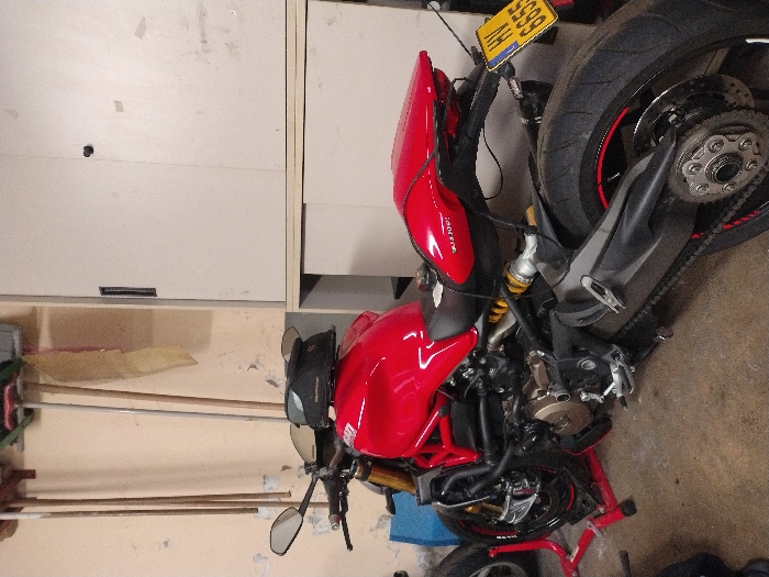 A red Ducati motorcycle, displaying Luxembourg license plate 'HV 6655' on a yellow background, is positioned on a red paddock stand within an indoor space in Fentange, Luxembourg. The motorcycle features a glossy red fuel tank and tail section, a black seat, and a visible engine with bronze-colored components. A black motorcycle helmet rests on the fuel tank. Gold-colored front suspension forks and a rear shock absorber with a yellow spring are present. The rear tire has a red stripe on its rim. The background on the left consists of a light-colored, possibly beige or tan, wall with visible wear and stains, from which several long-handled cleaning tools, including brooms and mops, are hanging vertically. To the right, two stacked light-grey metal storage cabinets are visible; the upper cabinet has two doors, one with a vertical handle, the other with a small black knob, while the lower cabinet appears to be a sliding-door unit. The floor is a dark-grey concrete surface.