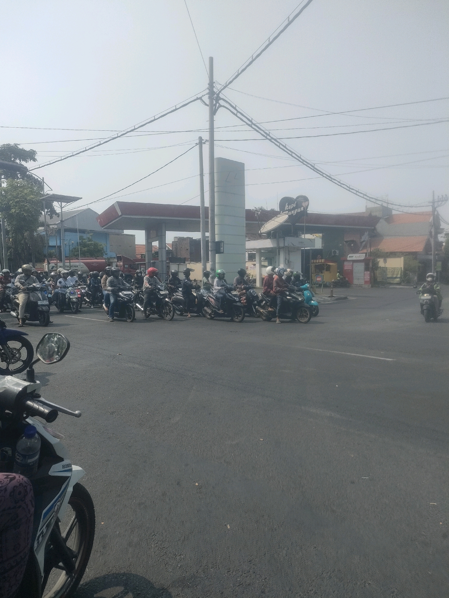 This image depicts a busy street scene in Surabaya, Indonesia, likely near a Pertamina gas station based on the signage and the presence of a large red fuel tanker in the background. The foreground shows a section of asphalt road with a white and blue Honda motorcycle on the left side, partially obscuring the view. The rider's leg, clad in patterned fabric, is visible. The motorcycle has a blue plastic water bottle strapped to it.
The mid-ground is dominated by a long line of motorcycles, many with riders wearing helmets, waiting at what appears to be an intersection or a traffic light. The riders are a mix of men and women. Some are looking forward, while others are glancing to the side. The motorcycles are varied in make and model, with a few scooters visible among them.
In the background, a Pertamina gas station with red canopies is prominent. Above and behind the gas station, a building with a sign that says "kopi angan" (meaning "dream coffee" in Indonesian) is visible. Power lines crisscross the sky, typical of an urban environment. The sky is hazy, suggesting it might be a hot or polluted day. On the far right, a single motorcyclist is seen on the road, heading away from the camera. The overall impression is one of a typical, bustling traffic situation in a densely populated Indonesian city.