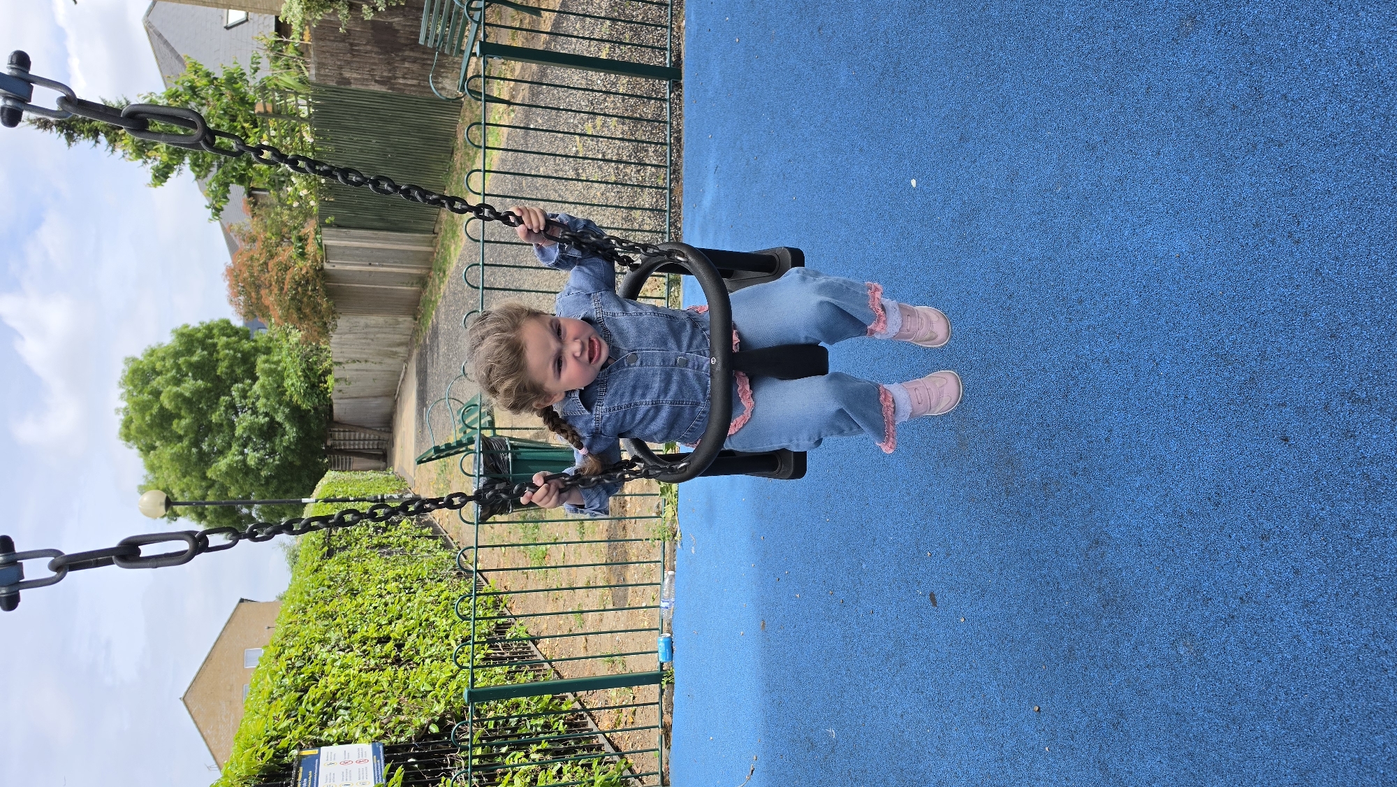 A young child wearing a denim outfit and pink shoes is on a swing set in a playground. The playground surface is blue. The swing set has black chains. The background features greenery, fences, and buildings. This photo appears to be taken in the United Kingdom, and could be in London based on the architecture and the presence of fencing commonly found in London playgrounds.