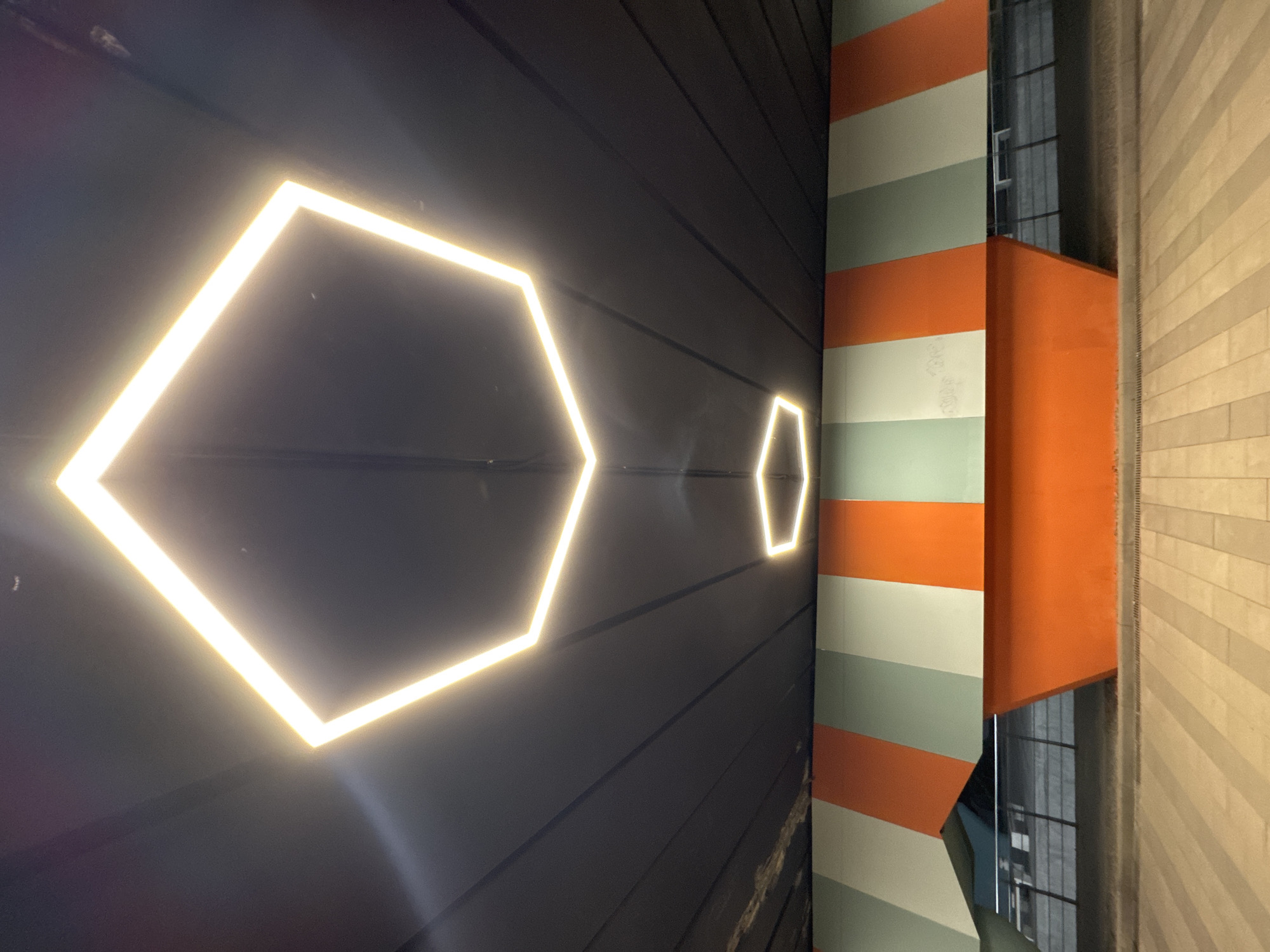 The image shows an underpass in Almaty, Kazakhstan. The ceiling of the underpass features two hexagonal LED light fixtures, one larger and one smaller, casting a warm glow. The walls of the underpass are decorated with a vibrant pattern of vertical stripes in orange, light green, and white. Below the striped section, a large, angular orange structure juts out. The ground level of the underpass is paved with light-colored rectangular tiles arranged in a diagonal pattern. In the background, behind the orange structure, there appears to be a metal railing and some fencing, suggesting a walkway or pedestrian area. The overall scene is modern and architectural, with a focus on geometric shapes and bold colors. There are no people visible in the image.