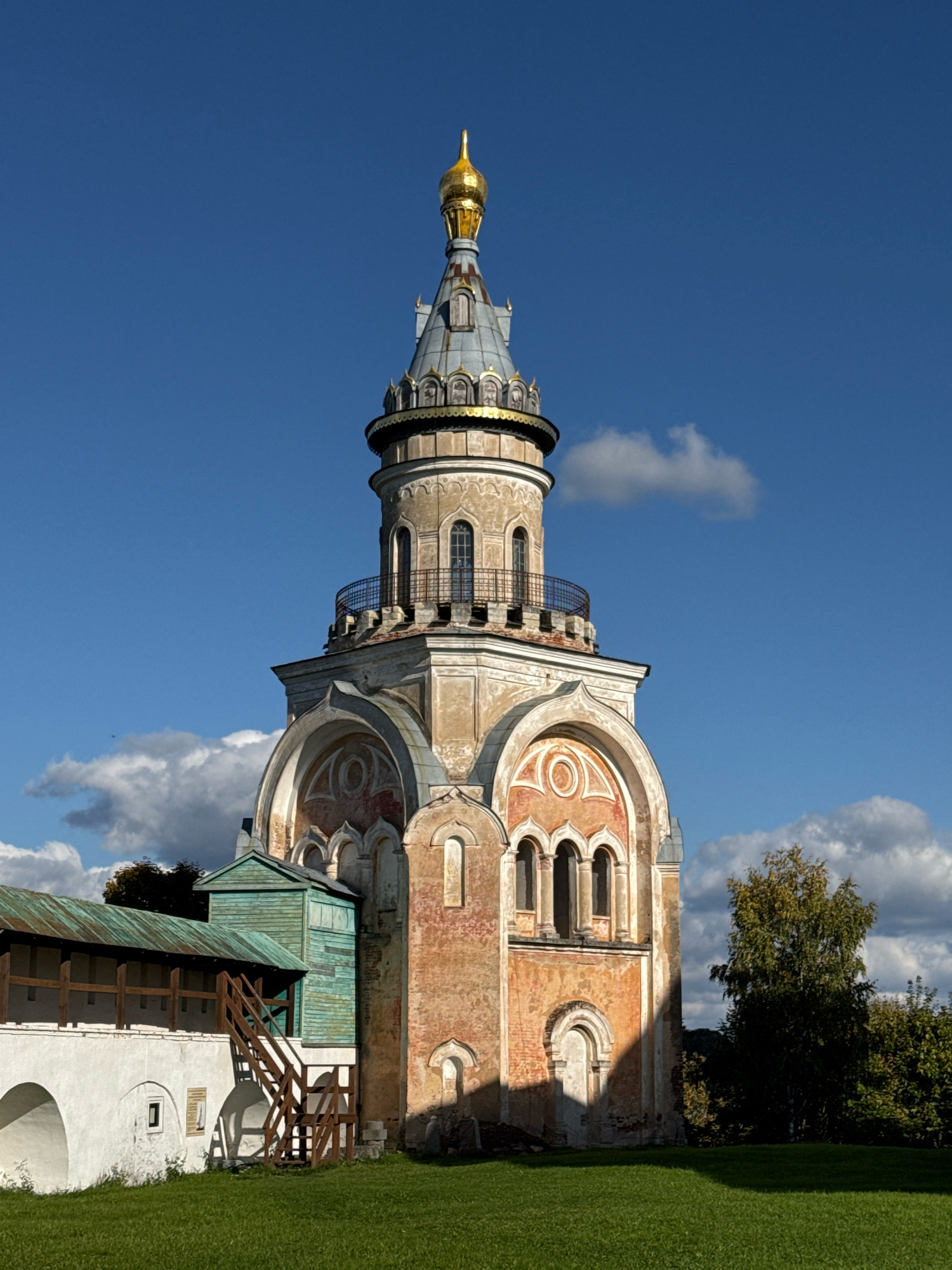 The image shows the Borisoglebsky Monastery bell tower in Torzhok, Russia. The bell tower is a tall, multi-tiered structure with a golden dome. It is made of brick and stone and has a weathered appearance, with some areas of discoloration and exposed brick. The tower is surrounded by a green lawn and trees. In the foreground, there is a wooden walkway with stairs leading up to a small, green building attached to the side of the monastery wall. The sky is blue with scattered white clouds. The sun is shining, casting shadows on the ground and the side of the bell tower.