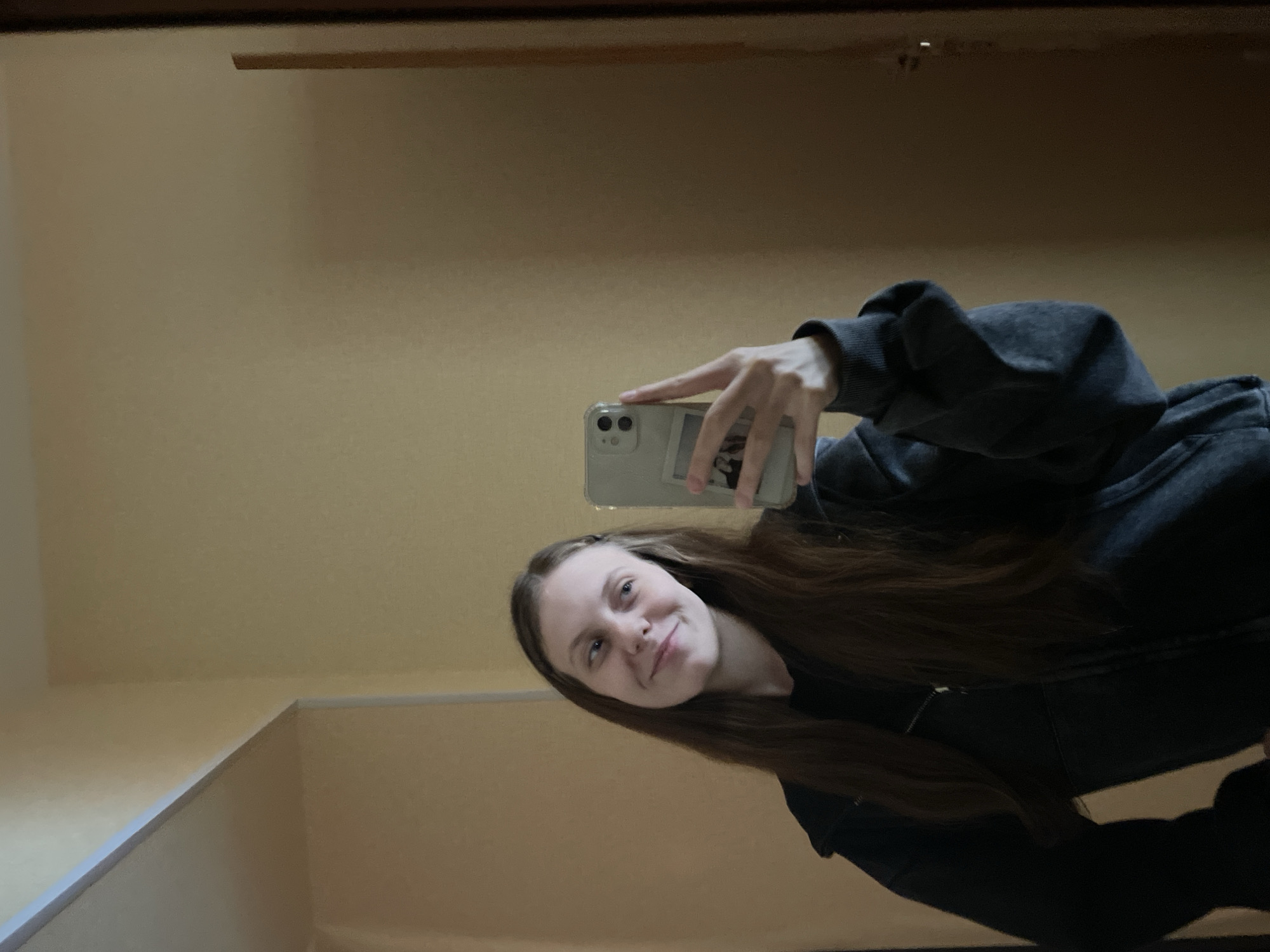 A young woman with long, brown hair is taking a selfie in a mirror. She is wearing a dark gray zip-up hoodie. The room appears to be a home interior with light yellow walls. The mirror reflects a doorway on the right side of the frame. The woman is smiling and looking slightly to her left. The lighting is soft and natural. There are no discernible landmarks or indicators to suggest the location is specifically in the city of Ufa, Russia.