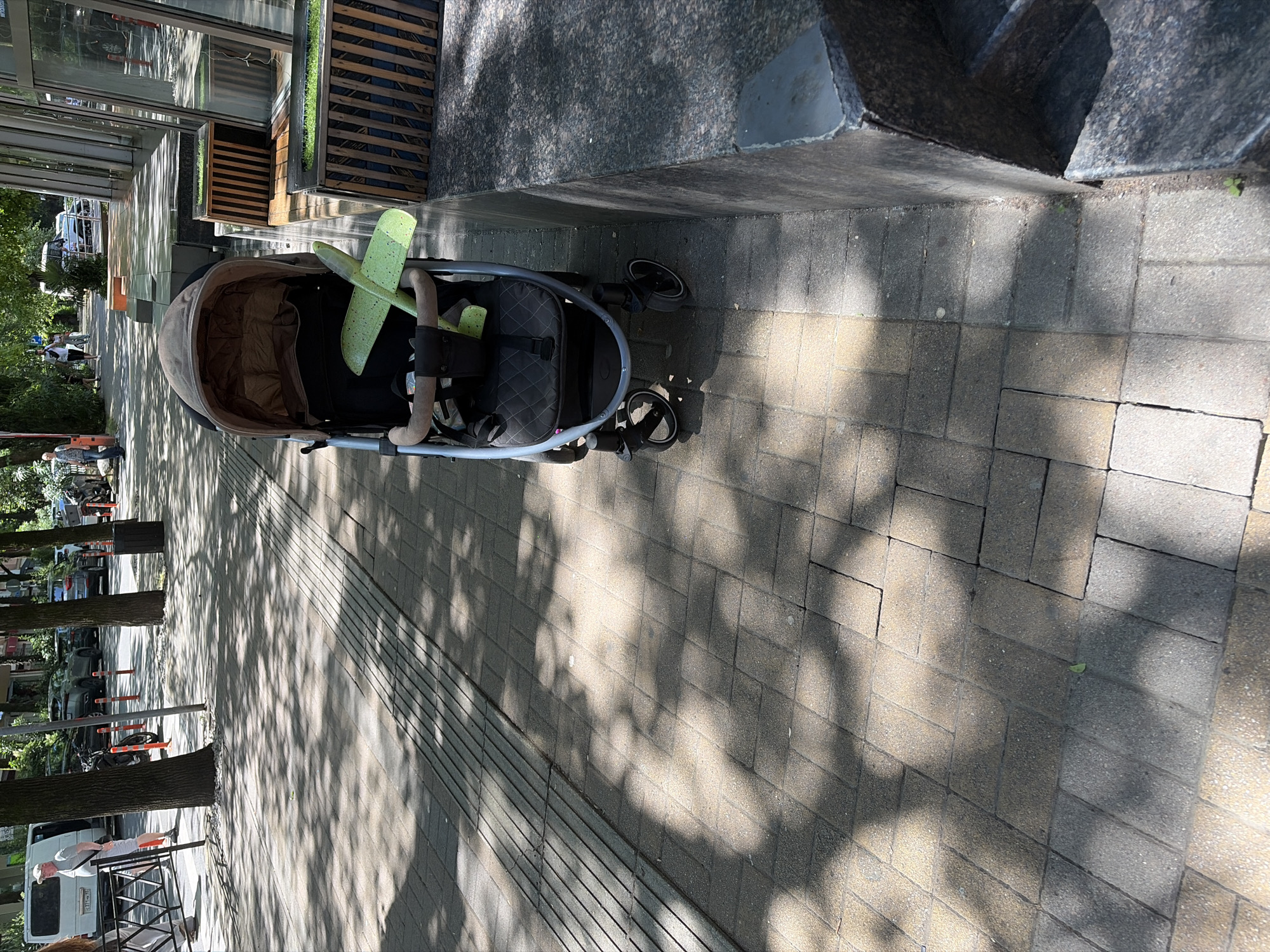 A stroller with a toy airplane on it sits on a sidewalk next to a building with large windows and planters. The sidewalk is made of rectangular pavers and is partially shaded by trees. In the background, there are cars and people walking on the street. The scene appears to be in a city.